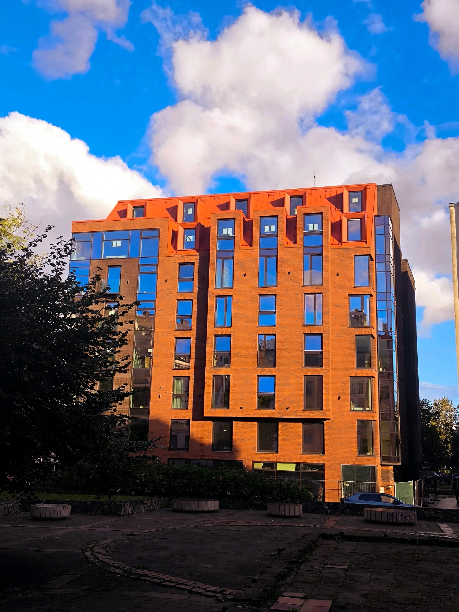 The image depicts a modern, multi-story brick building with an orange facade and a distinctive red roof featuring numerous dormer windows. The building has a prominent glass-covered section on its right side. A large, dark green tree partially obscures the left side of the building. The foreground shows a paved area with curved brick edging and several large, cylindrical planters. In the background, a silver car is parked, and there are indications of ongoing construction or landscaping work, including green fencing. The sky is a vibrant blue with scattered white clouds, suggesting fair weather. The scene appears to be set in a residential or commercial area. Although the exact location within Riga, Latvia, is not explicitly stated in the image, the architectural style and urban setting are consistent with contemporary developments in the city. There are no discernible people or specific actions taking place in the image.