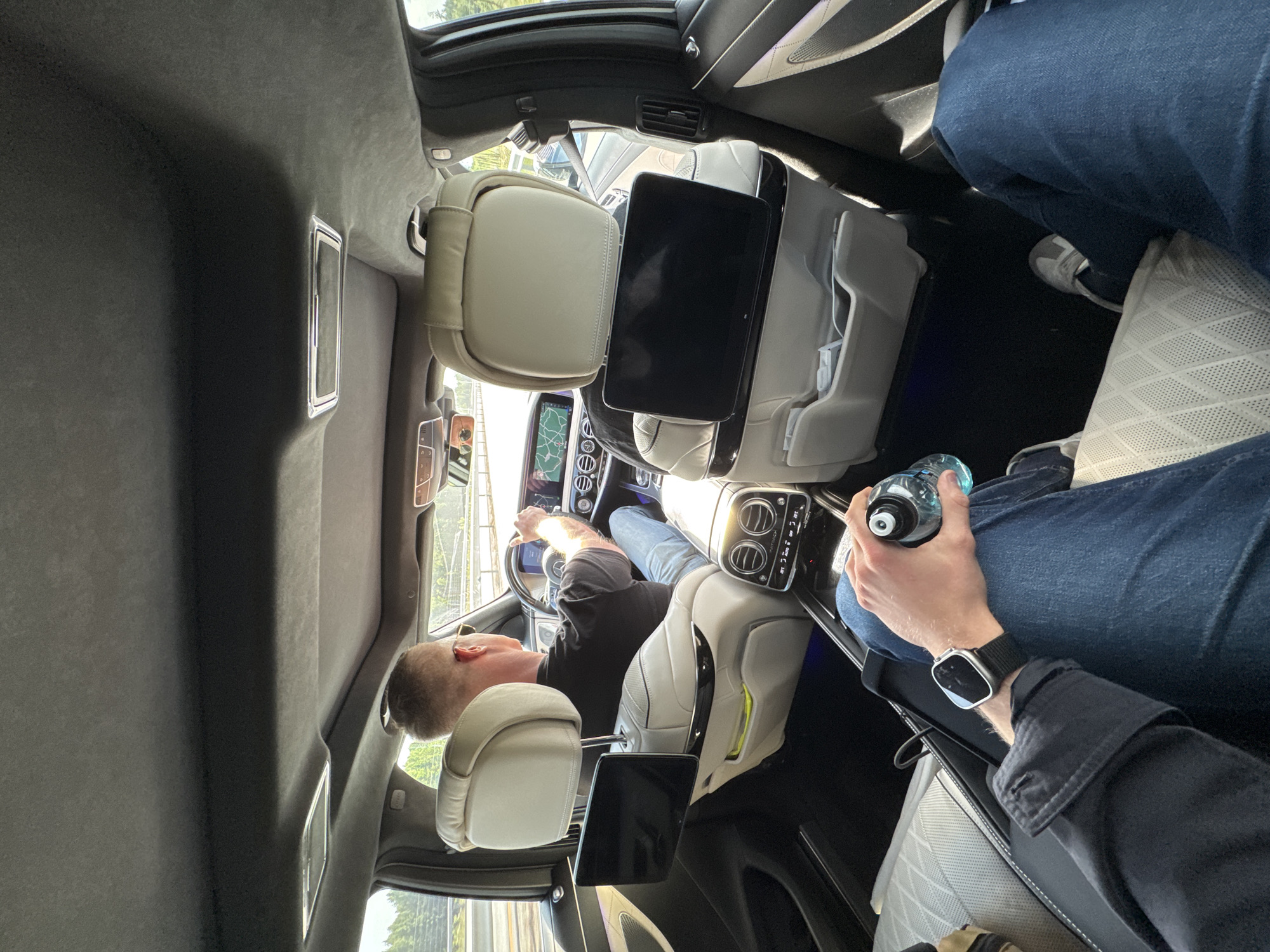 A person, presumably the driver, is positioned in the driver's seat of a car and appears to be actively driving. The car's interior, notably the seats and technological interfaces, suggests a luxury vehicle. A second person is in the rear passenger seat, holding a water bottle. The location is Ottestad, Norway, as indicated by the visual context.