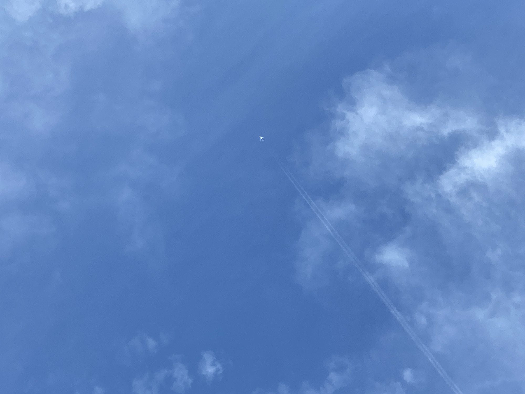 The image displays a clear blue sky, partially occupied by diffuse white clouds. A white airplane is visible in the upper-right quadrant, leaving a distinct double contrail that extends diagonally from the upper-right toward the lower-left corner of the frame. The scene is illuminated by natural daylight, observed from a location in the city of Unknown, Unknown.