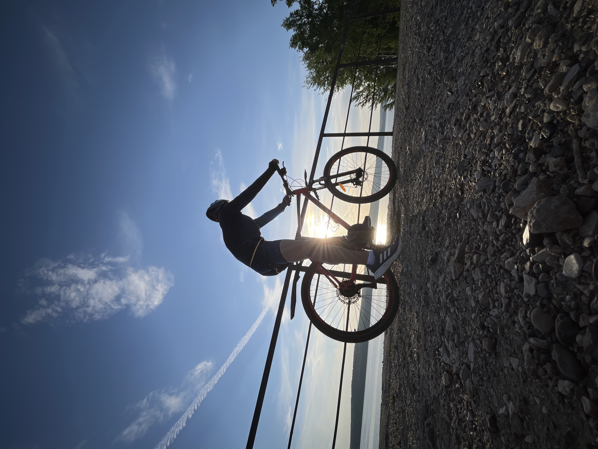 A person riding a bicycle near a railing next to a body of water is backlit by the sun in a low-angle shot. The bicycle is red. The person is wearing a dark long-sleeved shirt, light-colored shorts, and sneakers with three white stripes. A helmet is visible on their head. The railing has a metal top bar and vertical supports. The ground next to the railing is covered in small rocks. In the background, trees and a body of water are visible. The sky is blue with some scattered clouds and contrails. The location is Chaikovsky, Russia.