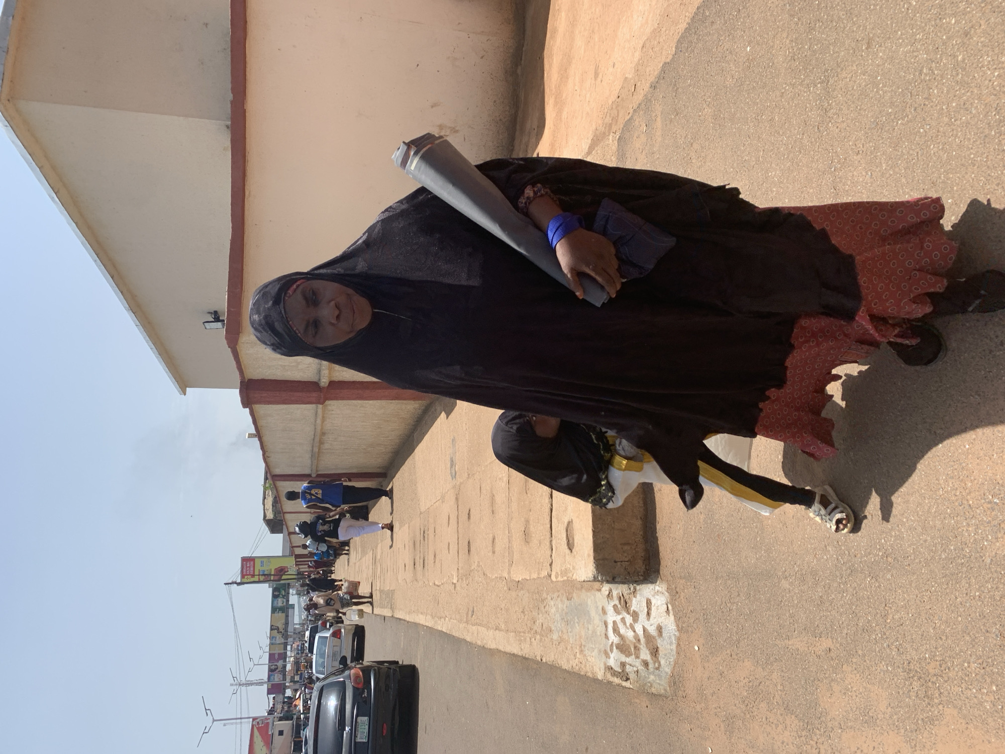 An adult woman and a child are walking on a sidewalk in Jos, Nigeria. The woman is wearing a dark colored hijab and dress. She is carrying a large rolled-up object in her right arm, and a blue bracelet is visible on her left wrist. The child is wearing a dark hijab and a white and yellow garment, with black leggings and white sandals. They are walking on a concrete sidewalk next to a street with cars and other pedestrians in the background. The buildings along the street appear to be commercial or residential. The sky is clear and the sun is bright, suggesting it is daytime.
