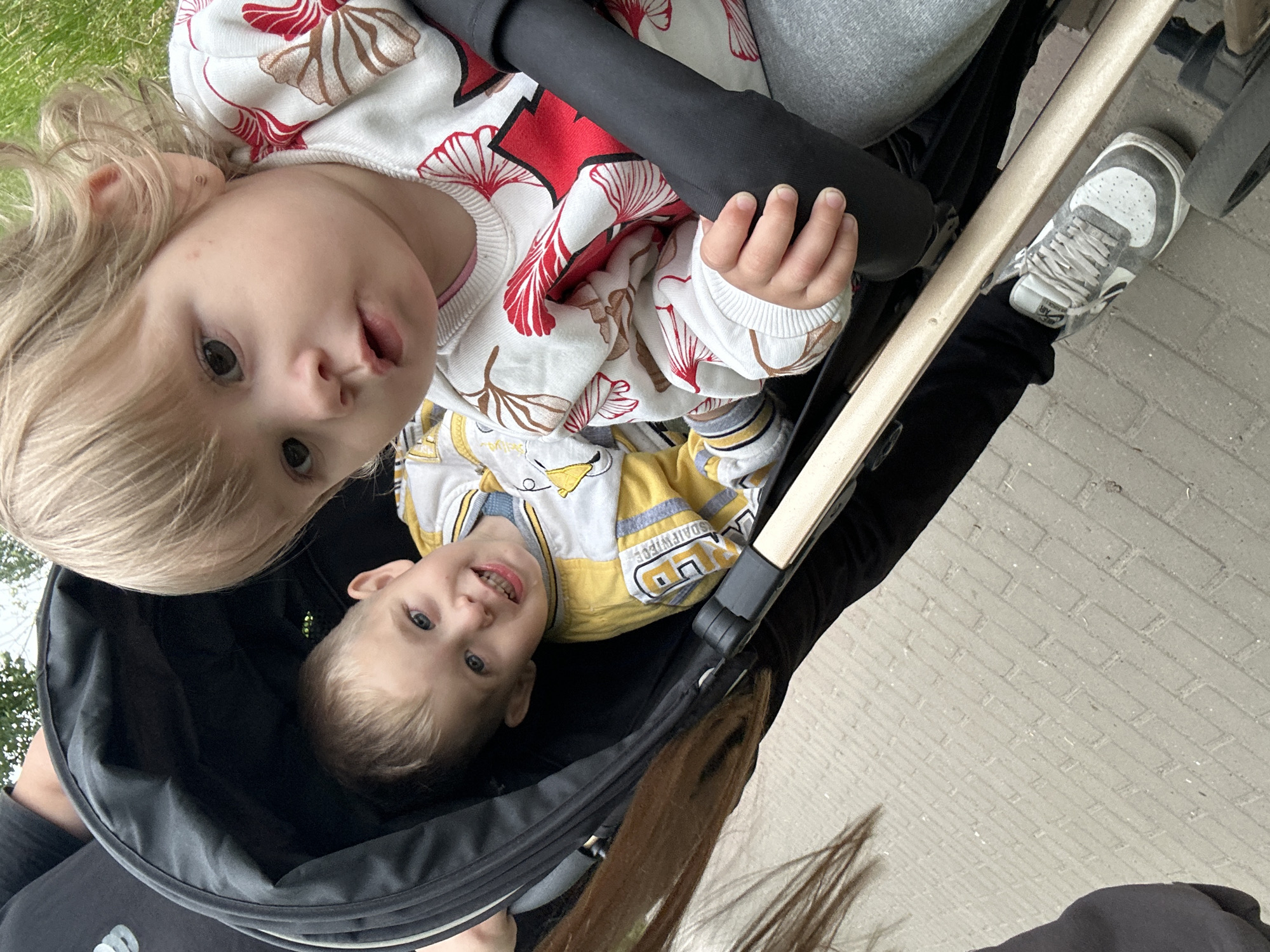 Two children are in a stroller on a brick walkway. The child on the left is a boy with short blonde hair and blue eyes, wearing a yellow and white striped shirt. The child on the right is a girl with blonde hair, wearing a white sweater with red and brown floral patterns. Her left hand is resting on the stroller's bar. A person's dark-colored leggings and a gray sneaker are visible in the foreground. The background shows greenery and a brick pathway, suggesting an outdoor, possibly park-like, setting in Chaikovsky, Russia.