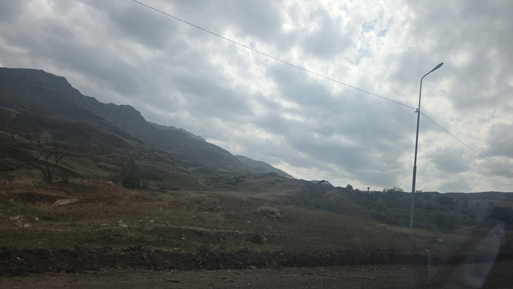 The image shows a panoramic view of a mountainous landscape, likely in the vicinity of Taldyk, Kyrgyzstan. The terrain is characterized by rolling hills and steep, rocky slopes covered in dry grass and sparse vegetation. A winding road or path is visible in the foreground, with a dark, earthy embankment. In the mid-ground, the hills rise up, with subtle terracing or agricultural patterns visible on some slopes, suggesting human activity. A few scattered trees and some small buildings or structures are discernible on the distant hillsides. A tall, slender streetlight stands prominently in the mid-right portion of the image, its pole silhouetted against a cloudy sky. A thin wire stretches across the frame, from near the streetlight towards the left. The sky is overcast with heavy, grey clouds, casting a subdued light over the scene. The overall impression is one of a rural, somewhat rugged, and remote natural environment.