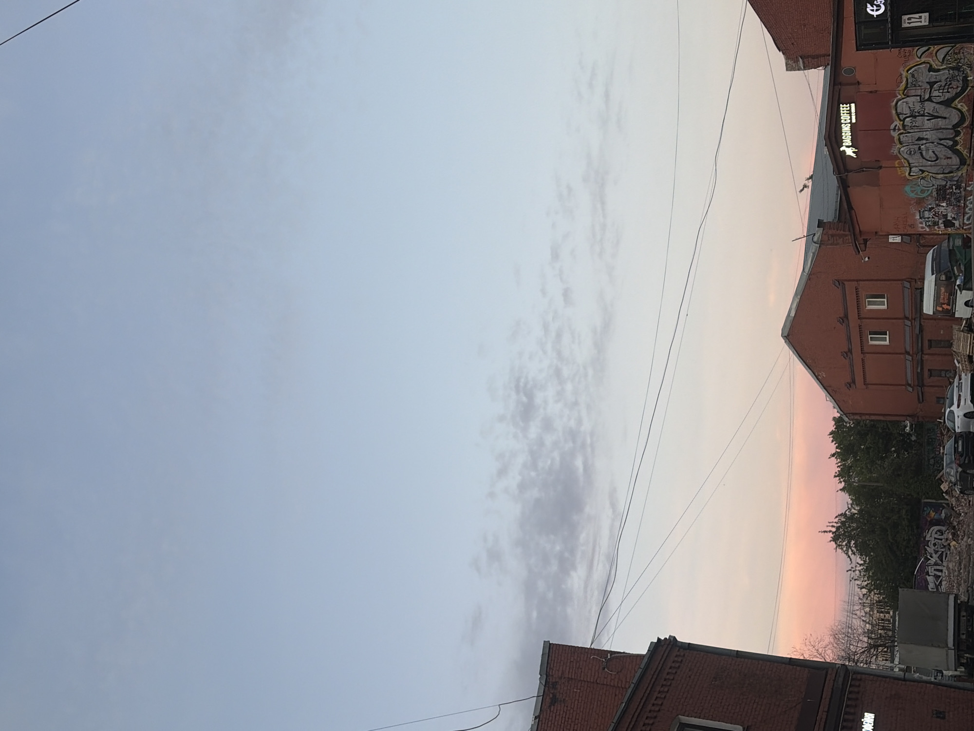 The image depicts a street scene in Saint Petersburg, Russia, likely during the late afternoon or early evening, given the soft, pastel colors in the sky. The sky is a pale blue with scattered wispy clouds, transitioning to a soft pink hue near the horizon, suggesting sunset.
In the foreground and mid-ground, brick buildings with orange and red facades are visible. Some buildings feature signs, including one that reads "BAGGINS COFFEE". The architecture appears to be industrial or utilitarian, with several windows and dark roofs. The buildings are adorned with graffiti, some of which is quite prominent and covers large sections of the walls.
The street level shows signs of construction or demolition, with piles of debris, including rubble and wooden pallets. Several vehicles are present: a white van, a dark-colored car, and a white car. A portion of a grey truck is also visible on the left side of the image. Overhead, numerous electrical wires crisscross the scene, a common feature in urban environments. Trees with bare branches, indicating a cooler season, are scattered between the buildings and behind the construction debris. The overall impression is one of an urban industrial area undergoing some form of transformation or renovation.