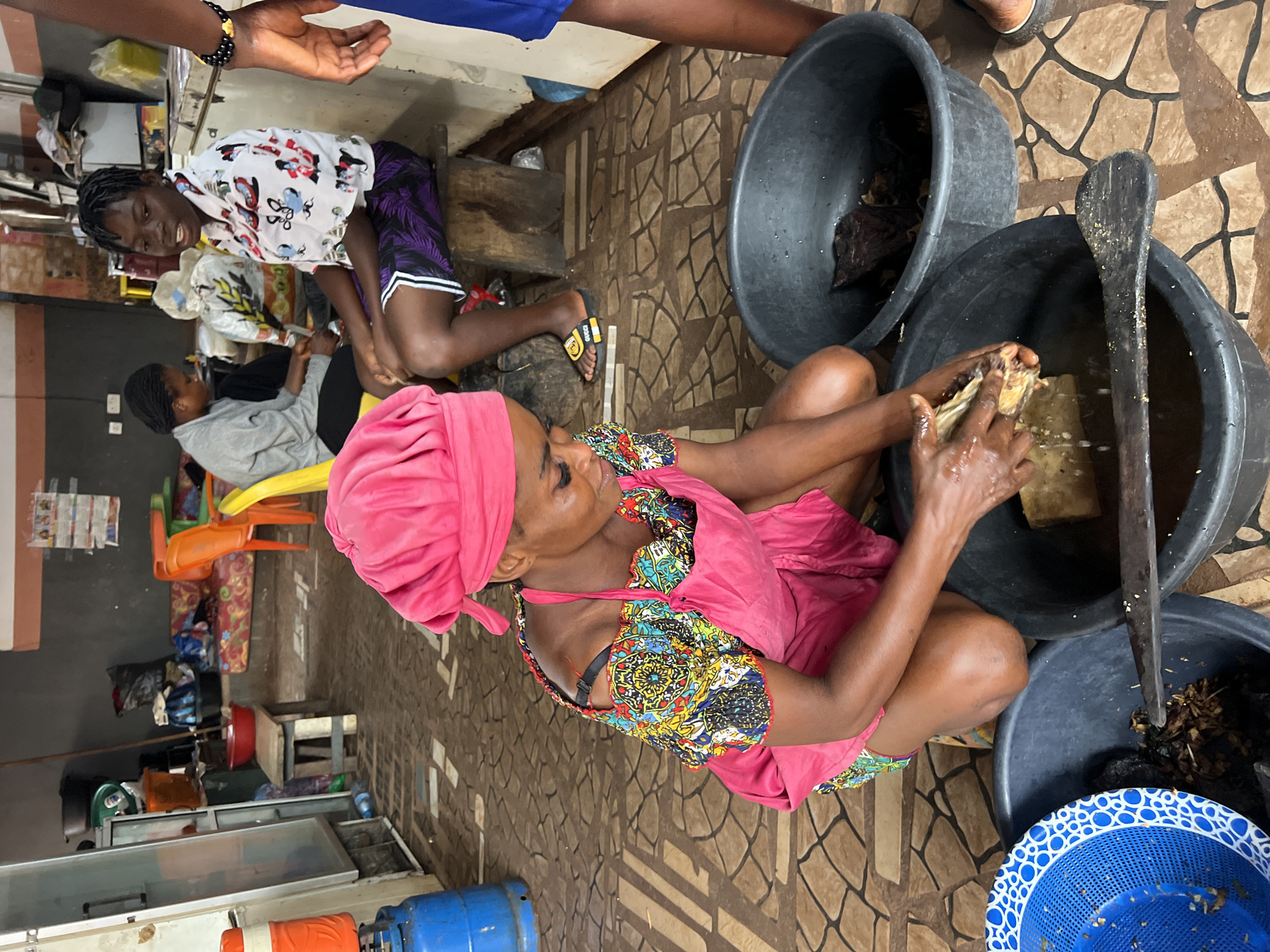 The image depicts a dimly lit indoor space in Nkpor, Nigeria, likely a market stall or food preparation area. A woman in a bright pink headscarf and apron is kneeling on the floor, her hands immersed in a large black basin filled with water and what appears to be fish or meat. She is actively working on a piece of food, which is partially submerged. Another black basin sits beside her, containing more processed food items. A blue colander with a white patterned rim is to her left, holding some food scraps. The floor is tiled with a geometric pattern of brown and beige squares. In the background, several other individuals are visible, including a young woman in a white shirt with colorful prints and purple shorts, sitting on a wooden stool. Another person in a gray sweater is seated further back. Various items are scattered around the space, including furniture like a sofa and chairs, containers, and what seems to be a gas cylinder. The overall impression is one of daily activity in a commercial or domestic setting focused on food processing.