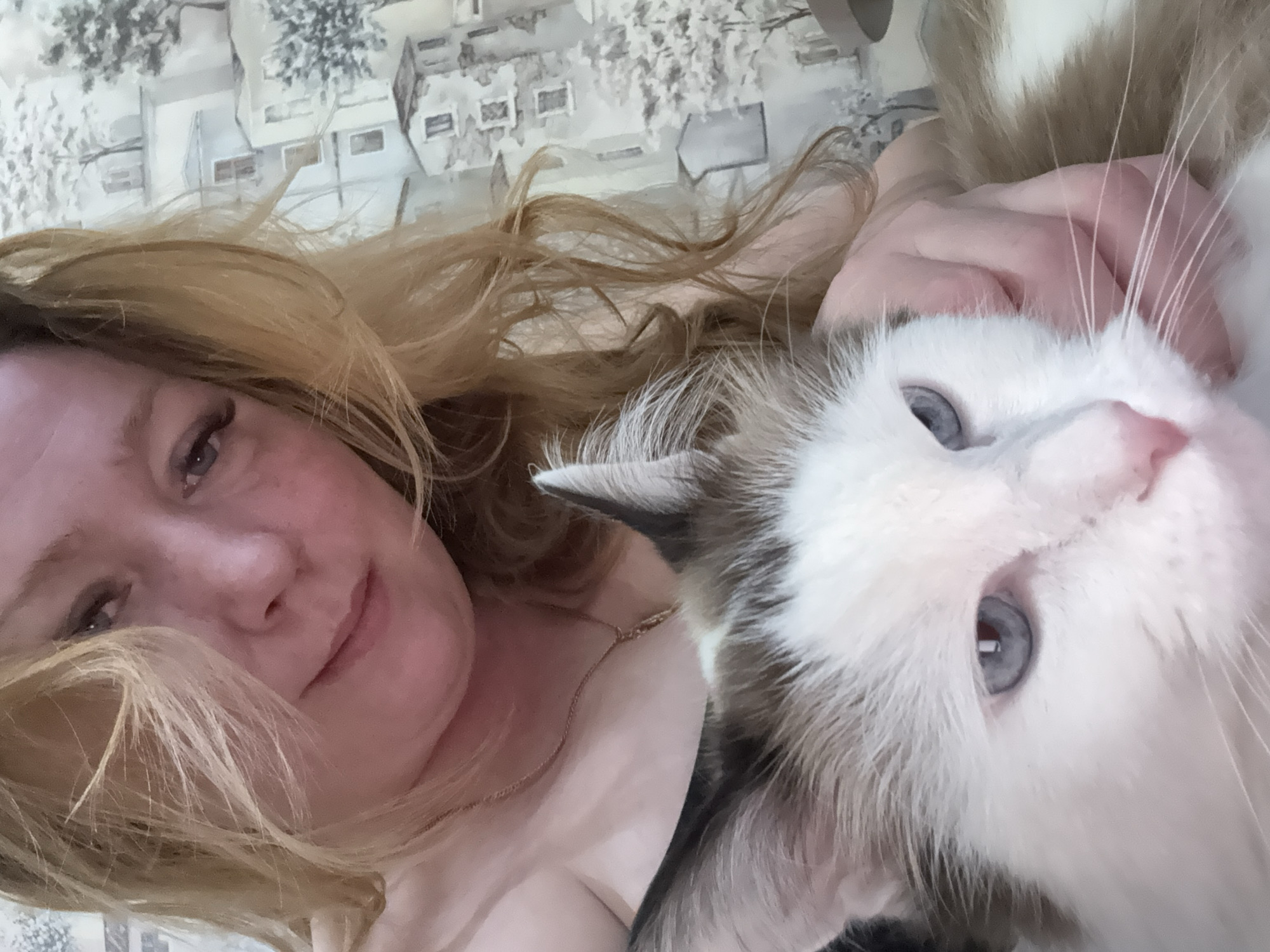 A woman with long, blonde hair is lying down, close to the camera, with a fluffy white and brown cat nestled against her. The cat has blue eyes. The woman is wearing a light-colored top and a delicate necklace. The background is a patterned wall or covering with a depiction of buildings. The image's location is not identifiable in the city of Izhevsk, Russia.