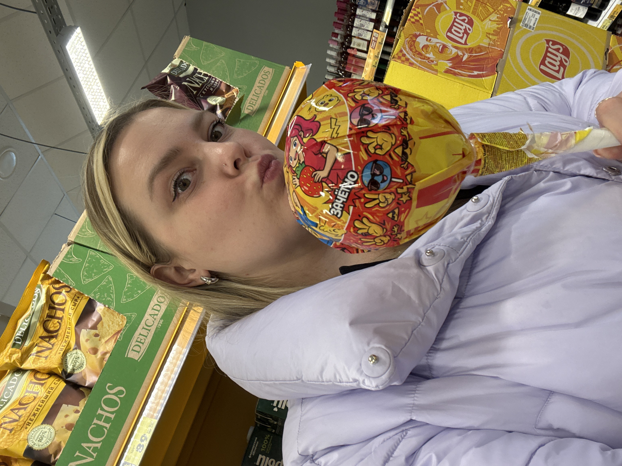 A young woman in a light purple puffer jacket is holding a large, wrapped lollipop in front of her face, pursing her lips as if kissing the candy. She is standing in what appears to be a grocery store aisle, with shelves filled with snack items and drinks behind her. On the left, there are several packages of "Delicados Nachos" with a green and yellow color scheme. Above the nachos, there is another package of "Delicados Nach." On the right side of the frame, there are boxes of "Lay's" potato chips. The lollipop itself is round and covered in yellow and red wrapping with cartoon characters and the word "ЗАЧЁТНО." The lighting in the store is bright and fluorescent. The setting is likely a store located in Chaikovsky, Russia, due to the Russian text on the lollipop.