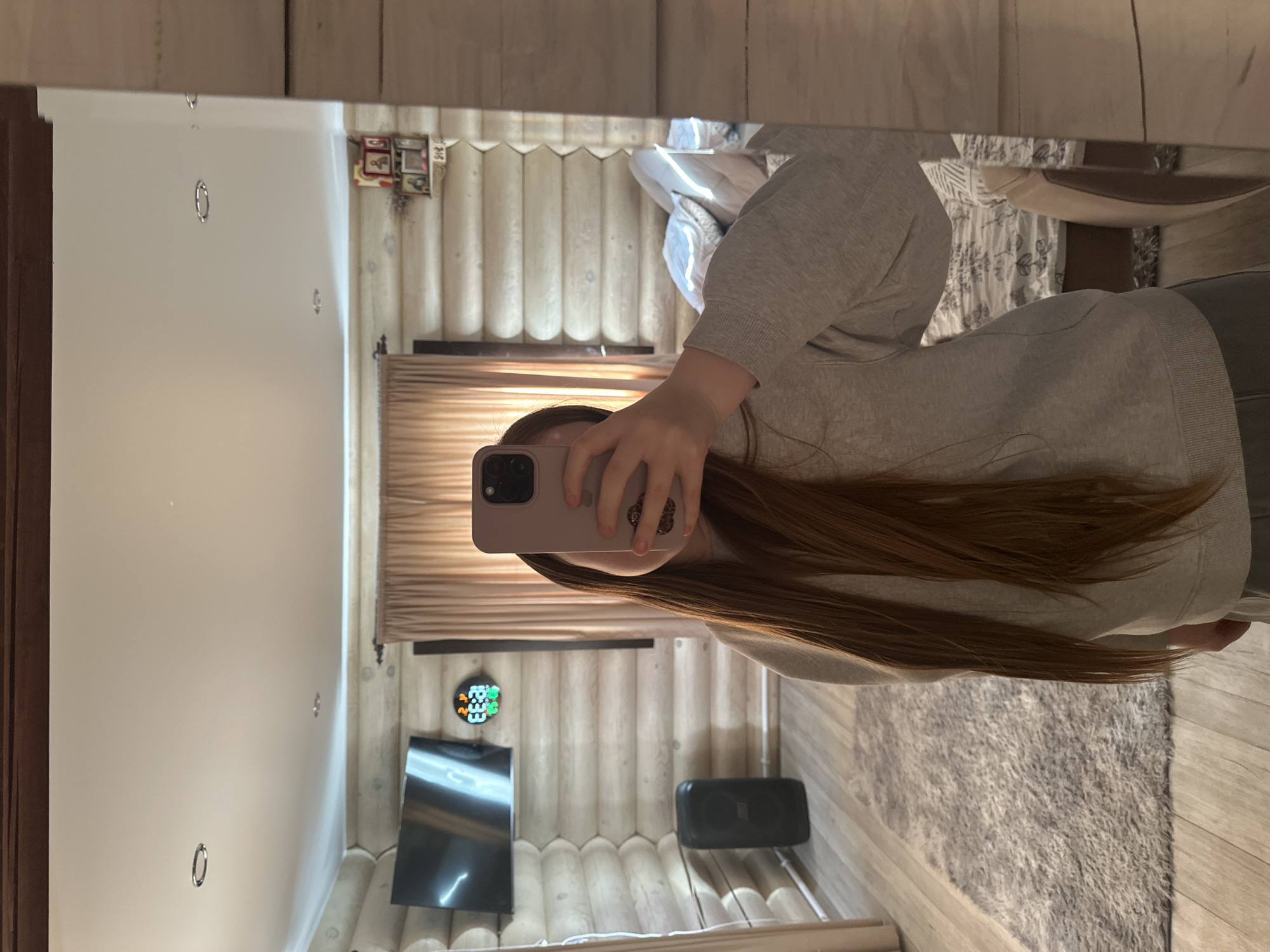 The image appears to be a mirror selfie taken inside a room constructed with light-colored log walls, possibly in a house or cabin. The room is furnished with a bed, a TV, and a speaker. A woman is holding up a phone to take the selfie, obscuring her face. Her hand is visible holding the phone and long brown hair extends from behind the phone. A rug is visible on the floor. Small pictures are visible hung on the wall. The exact location in Ольховка, Russia, cannot be determined from the image.