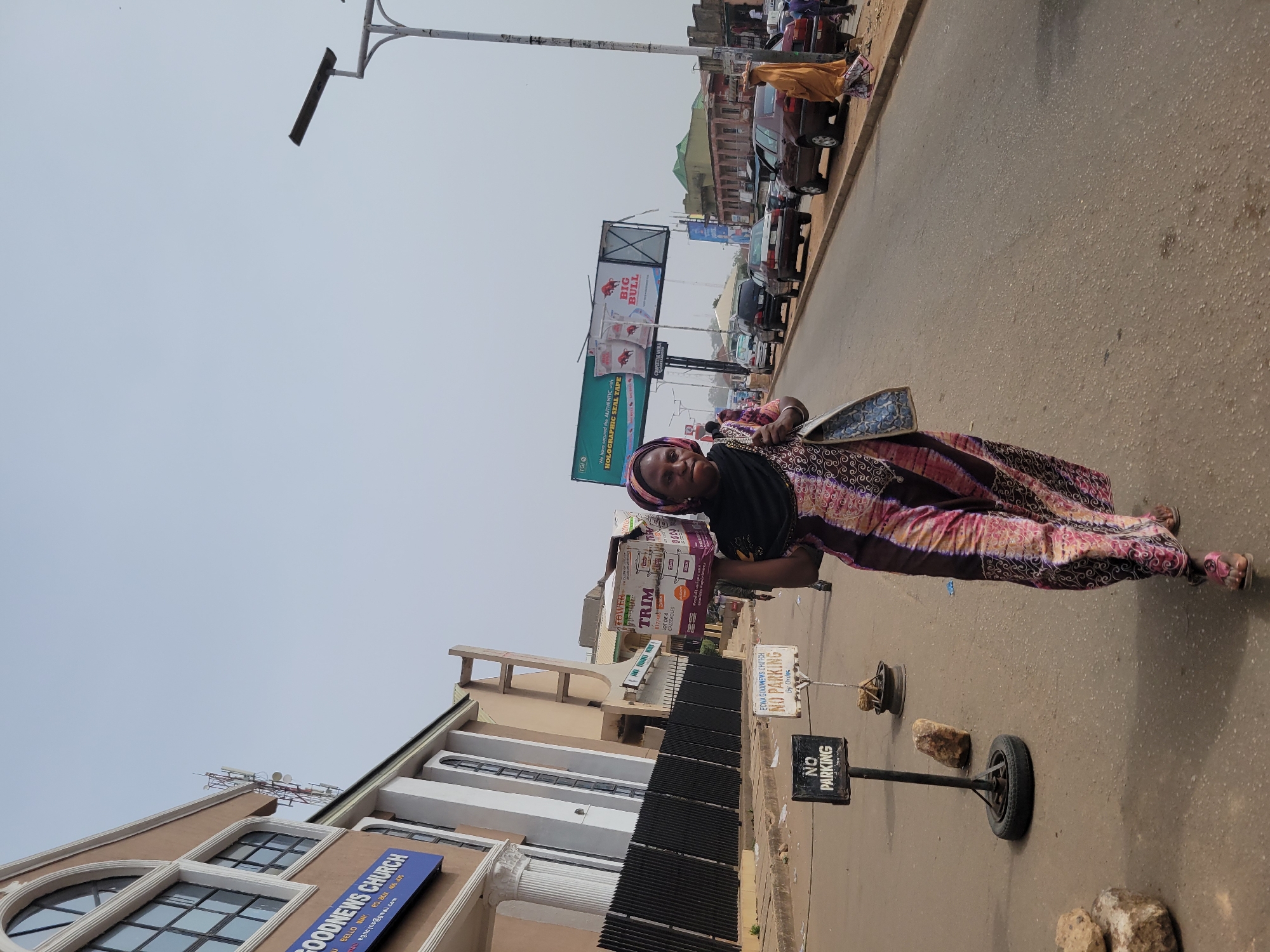 The image presents a daytime street scene in Jos, Nigeria, under a bright, overcast sky. In the foreground, on the right side of the street, a woman is captured looking directly at the camera. She wears a black head covering, a patterned dress in shades of brown, pink, and white, and sandals, carrying a bag with a geometric design.
The asphalt street extends into the background, where several cars are parked along the right side. On the left, a prominent light-brown building with arched and rectangular windows is visible. A blue sign on this building identifies it as "GOODNEWS CHURCH," with additional text "BELLO WAY, PO BOX 40%, JOS" and "mail egnc.jos@gmail.com." Further along the street, another sign confirms it as "ECWA GOODNEWS CHURCH."
Near the church, a black metal fence stands alongside two "NO PARKING" signs, one of which specifies "By Ordes." These signs are anchored by unconventional bases, including rocks and an old tire rim. In the mid-ground, several advertising billboards are visible. One prominent billboard features "BIG BULL," while another promotes "TRIM CASSEROLES" and mentions "LOT DE 4" and "We have secured the AUTHENTIC with HOLOGRAPHIC SEAL TAPE." A utility pole with a streetlight extends into the upper frame. The scene portrays a lively urban environment.