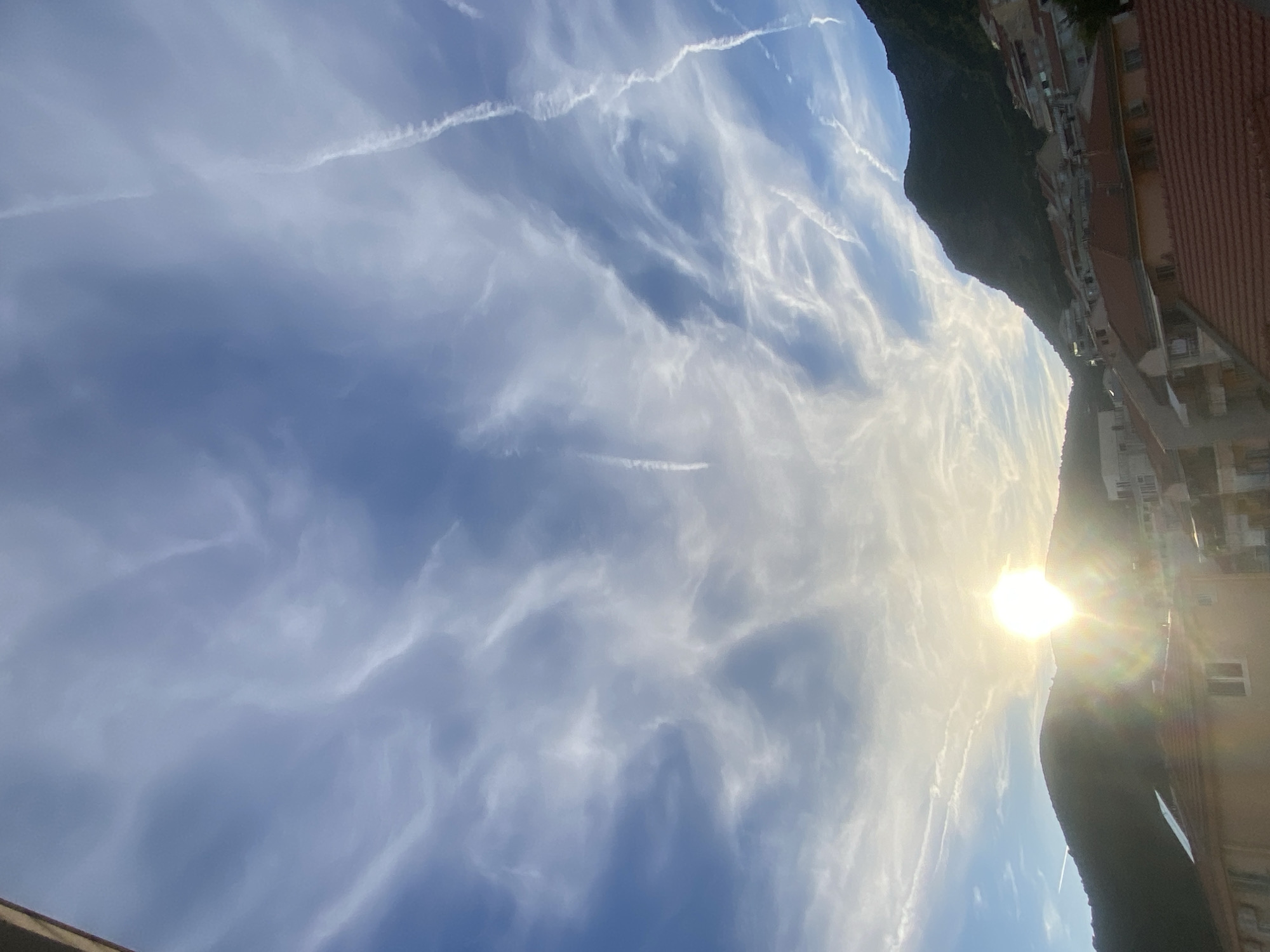 The image depicts a view of the sky over a coastal town in Budva, Montenegro, likely during sunrise or sunset due to the presence of the sun near the horizon. The sky is a vibrant blue, filled with wispy white clouds that create a dynamic and textured appearance. Several contrails from aircraft are visible, streaking across the sky.
In the foreground and midground, a hillside is covered with buildings, suggesting a densely populated area. The architecture appears to be typical of a Mediterranean coastal town, with terracotta roofs visible on many structures. The buildings are nestled into the slopes of the hills, with some showing signs of age and wear.
The hills themselves are dark and silhouetted against the bright sky, their contours suggesting a rugged natural landscape. The sun is a prominent feature, casting a bright, golden glow and creating a lens flare effect that illuminates the scene. The overall impression is one of natural beauty combined with human habitation in a scenic coastal environment. There are no people or specific actions depicted in the image.