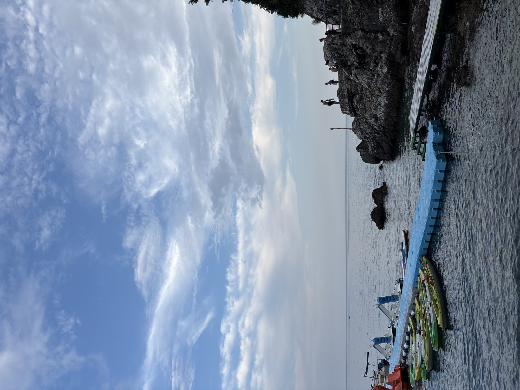 This media file captures a scenic view of a rocky coastline with people cliff jumping into the ocean. The scene is set during what appears to be daytime, with a clear blue sky adorned with wispy white clouds. The water is calm and reflects the sky.
The primary subjects are a group of people, some of whom are actively engaged in cliff jumping from a rocky outcrop. Others are visible on a metal railing at the top of the cliff, observing the activity. In the foreground, a blue modular floating dock is visible, with several kayaks and other small boats docked alongside it. A wooden pier extends from the rocks towards the floating dock. The overall atmosphere suggests a recreational and adventurous setting.
Notable details include the dynamic poses of the cliff jumpers, some mid-air, conveying a sense of thrill and excitement. The rocky terrain is rugged and imposing, contrasting with the serene expanse of the sea and sky. The presence of kayaks and boats indicates a potential for water sports and exploration.
The location context is Budva, Montenegro, which aligns with the visual cues of a coastal area with rocky cliffs and clear waters. The time of day is likely late afternoon or early evening, given the soft lighting and the angle of the sun. There is no visible text within the media file.