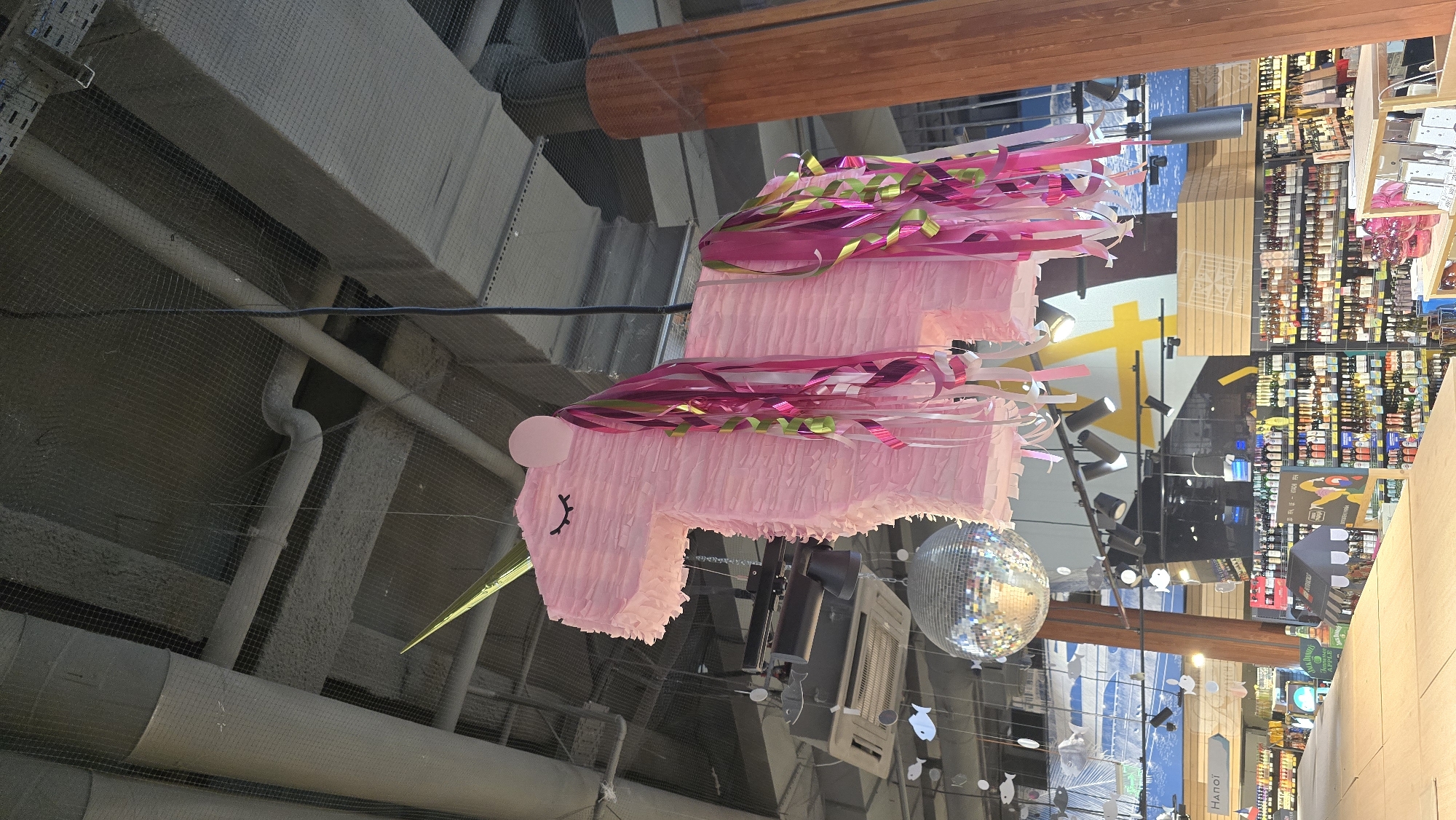 A large, pink unicorn-shaped piñata, adorned with vibrant pink, fuchsia, green, and white streamers forming its mane and tail, hangs prominently from a high ceiling. The piñata features a pointed green horn, a small pink ear, and black lines indicating closed eyes.
Below and to the right of the piñata, a reflective disco ball is suspended, casting light patterns. The overhead structure reveals an industrial-style ceiling with visible gray ventilation ducts, pipes, and metal racks, partially covered by a black mesh safety net. A large wooden beam runs across the upper part of the frame.
In the background, a brightly lit retail environment is visible, featuring numerous shelves stocked with various products, suggesting a supermarket or a large store, possibly a liquor aisle. Several track lights illuminate the merchandise. A large yellow graphic element is present on a wall, and small, white, fish-shaped decorations hang on wires in the mid-ground.
Partially visible text "Hanoi" appears on a sign or display towards the bottom right of the background. While the provided context suggests the location is Kyiv, Ukraine, this text indicates a different city. No people are visible, and the indoor setting means the time of day and weather are not discernible.