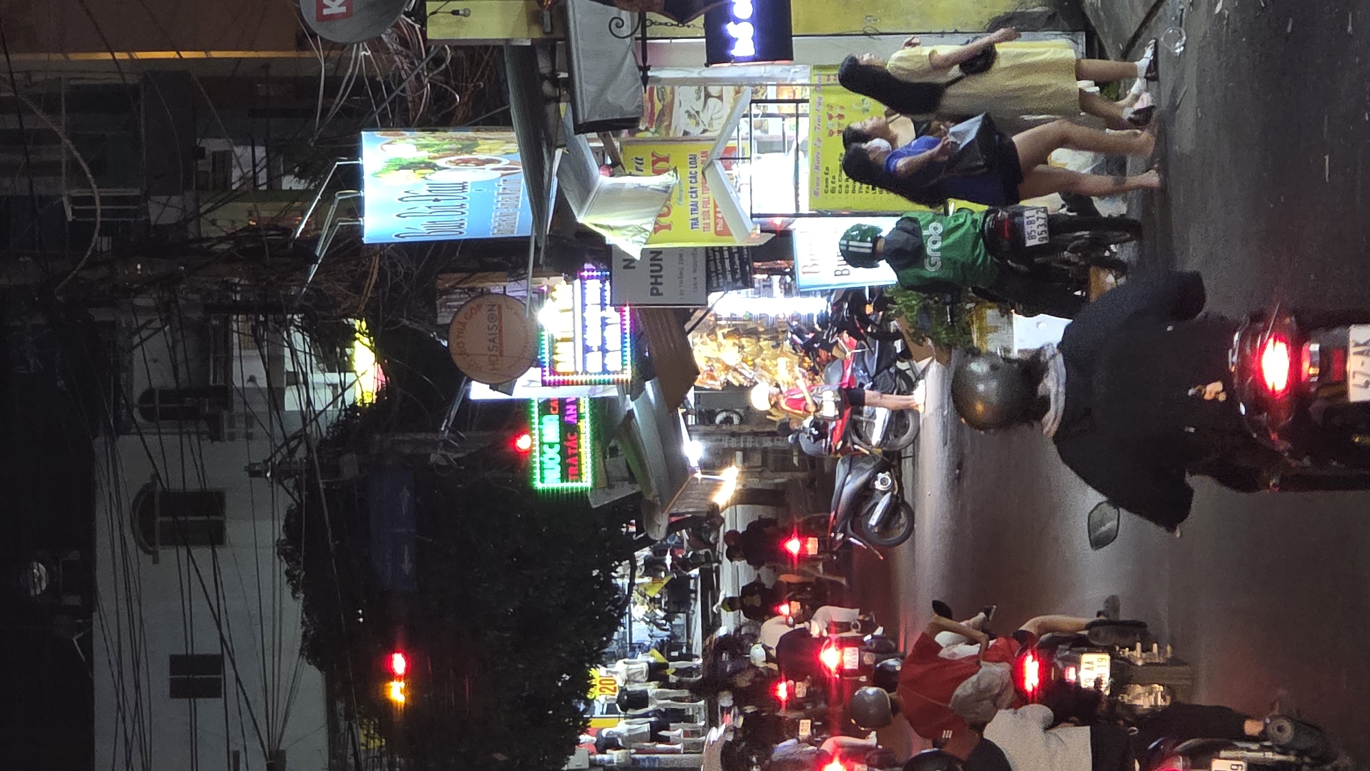 This is a vibrant, nighttime street scene in Bắc Nha Trang, Vietnam, bustling with activity. The image depicts a dense urban environment with numerous motorbikes, pedestrians, and brightly lit commercial signs.
In the foreground and midground, a busy street is filled with motorbikes, many stopped or moving slowly, indicated by the illuminated red tail lights. Several individuals are on their motorbikes, some wearing helmets, suggesting traffic or waiting at a light. A person wearing a distinctive green Grab delivery uniform is visible on a motorbike. On the right side of the street, two women are standing on the sidewalk, one in a yellow dress and the other in a blue top, both appearing barefoot.
The background reveals a series of multi-story buildings housing various businesses. The storefronts are adorned with numerous illuminated signs and advertisements in Vietnamese, casting colorful glows onto the street. Above the street, a dense tangle of electrical wires crisscrosses overhead, a common sight in many Southeast Asian cities. A satellite dish marked with "K" is visible high up.
Visible text on the signs includes "NƯỚC MÍA CÁ TRÁI TÁC AN" (likely referring to sugarcane juice and fruits), "HOA SAISON" (a brand or shop name), "TRÀ TRÁI CÂY CÁC LOẠI" (various fruit teas), "TRÀ SỮA FULL TOP" (full topping milk tea), "Cam Ép" (orange juice), and "PHUN". A specific date "2019" is also seen on a sign, possibly an opening year. A motorbike license plate displays "85-81 953.72". The overall impression is a lively commercial street after dark.