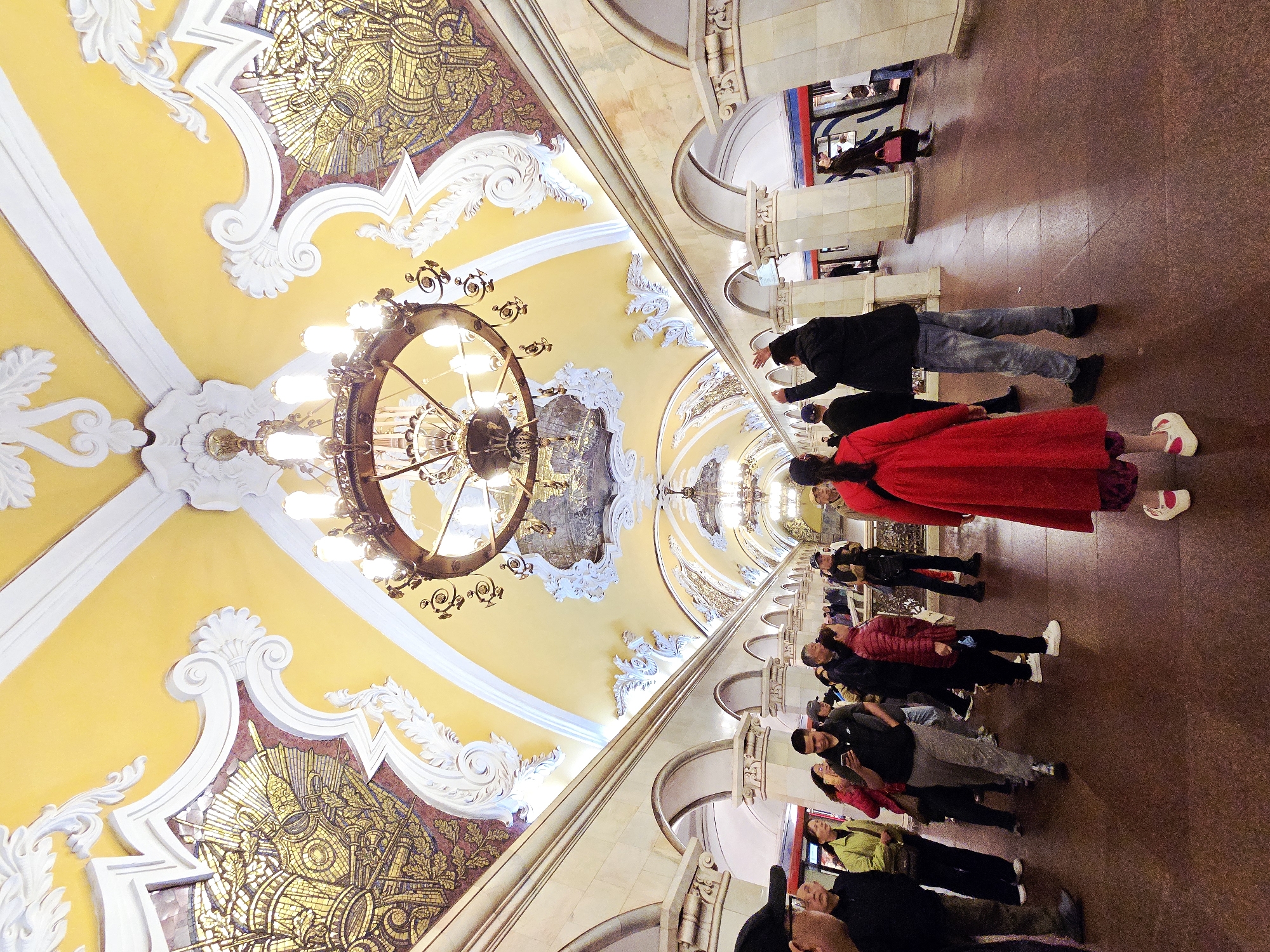 The image depicts a view inside a Moscow Metro station in Russia. The ceiling is adorned with intricate, ornate plasterwork, gold mosaics, and a large, circular chandelier. People are walking along the platform. Many are looking up at the ceiling. Some are taking photos. The architecture includes arches and pillars along the platform edge. A train platform is seen with a train waiting for people.