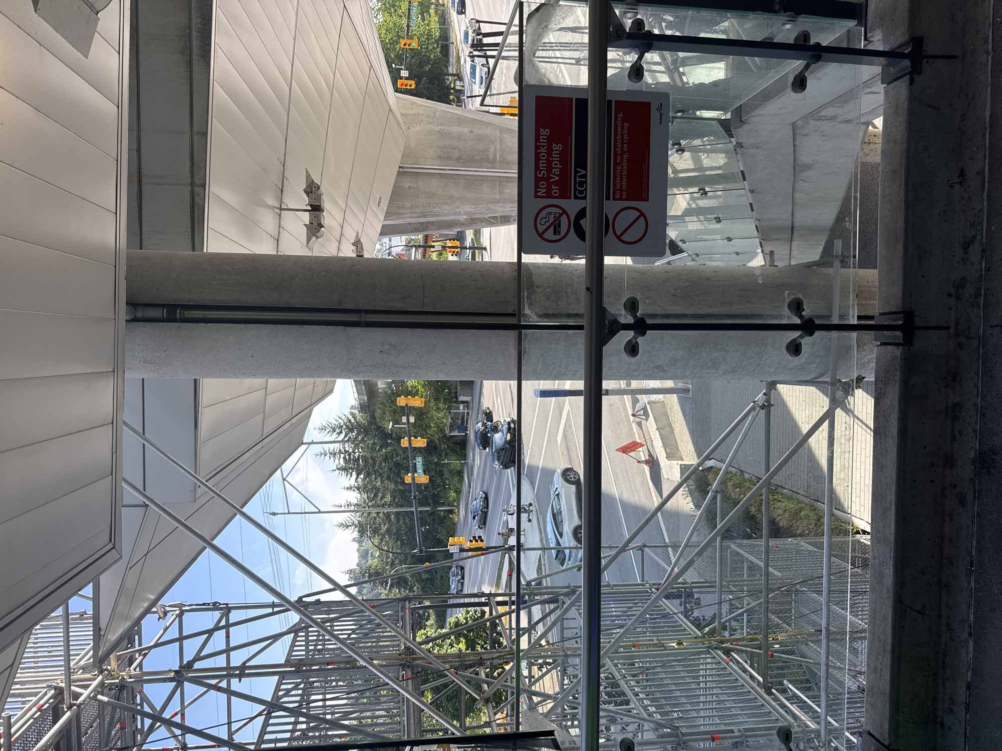The image shows a view of the Gilmore Station SkyTrain station in Burnaby, Canada, with a focus on the infrastructure of the station and the surrounding urban environment. In the foreground, there is a glass railing with metal fittings, suggesting an elevated viewpoint. To the left, a large scaffolding structure dominates the frame, indicating ongoing construction or maintenance work. Through the scaffolding, a street scene is visible with several cars driving on a road. Traffic lights are present, and one intersection has a "GILMORE" street sign.
Further into the background, a line of evergreen trees borders the road, and beyond that, more of the SkyTrain station's elevated structure is visible, including its white, angled roof. The sky is blue with some scattered clouds.
Two signs are prominently displayed on a concrete pillar. The top sign, in red and white, reads "No Smoking or Vaping" and also indicates "CCTV". Below this, another red and white sign lists prohibitions: "No loitering, no skateboarding, no rollerblading, no cycling." The bottom of this sign has the "TransLink" logo.
The overall impression is one of a functional public transit hub within a developed urban area, with elements of construction impacting the immediate view. The lighting suggests it is daytime, likely late afternoon due to the angle of the shadows. There are no individuals clearly visible in close proximity to the viewer.