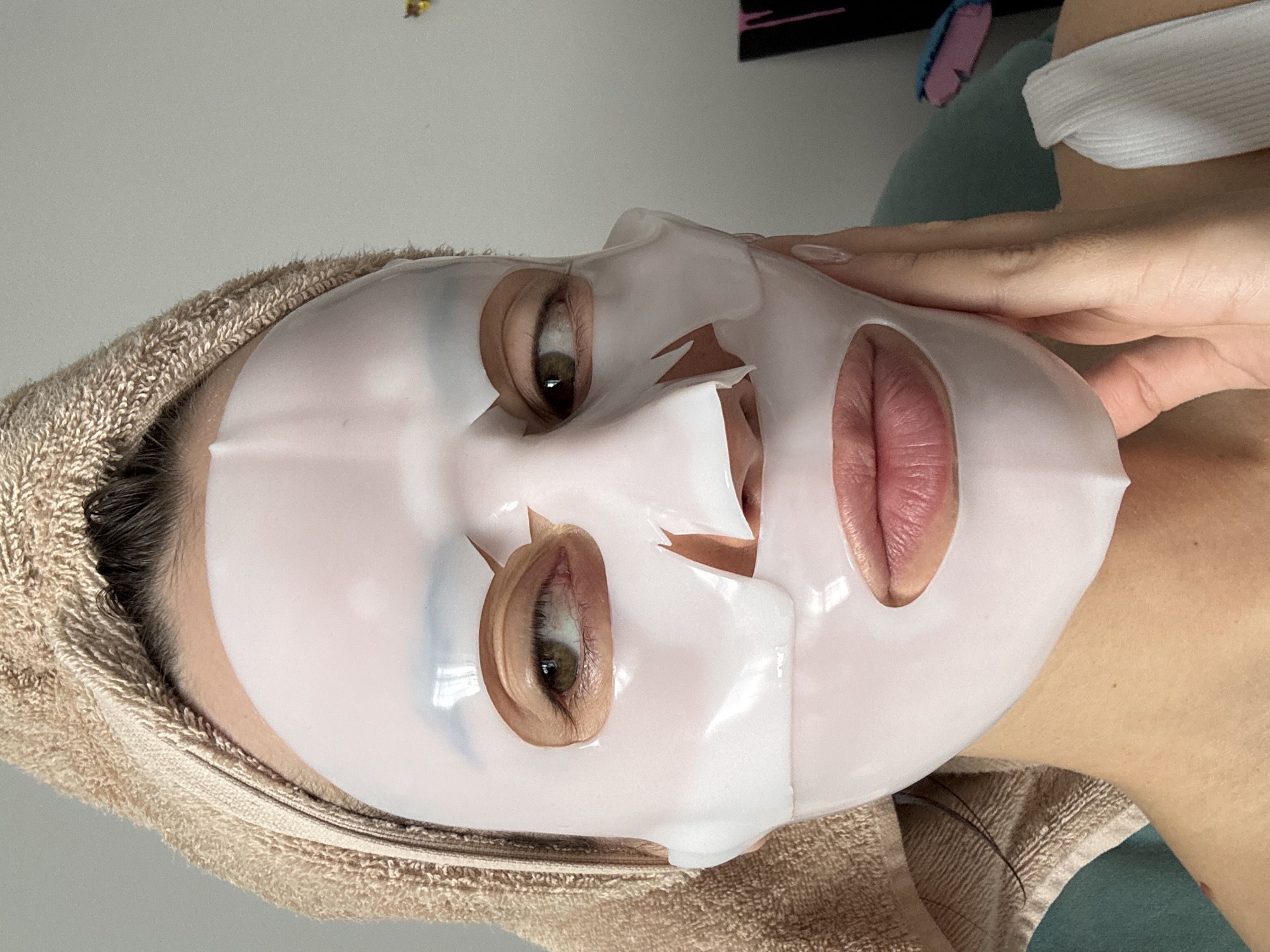 A person is lying down, facing the camera, wearing a white sheet mask and a tan towel wrapped around their head. The mask has cutouts for the eyes, nose, and mouth. A hand is visible on the right side of the frame, holding the mask in place. The person is wearing a white top with a visible strap. The background appears to be a combination of a white wall and green fabric. The location in the city of Krasnoyarsk, Russia cannot be determined from the image.