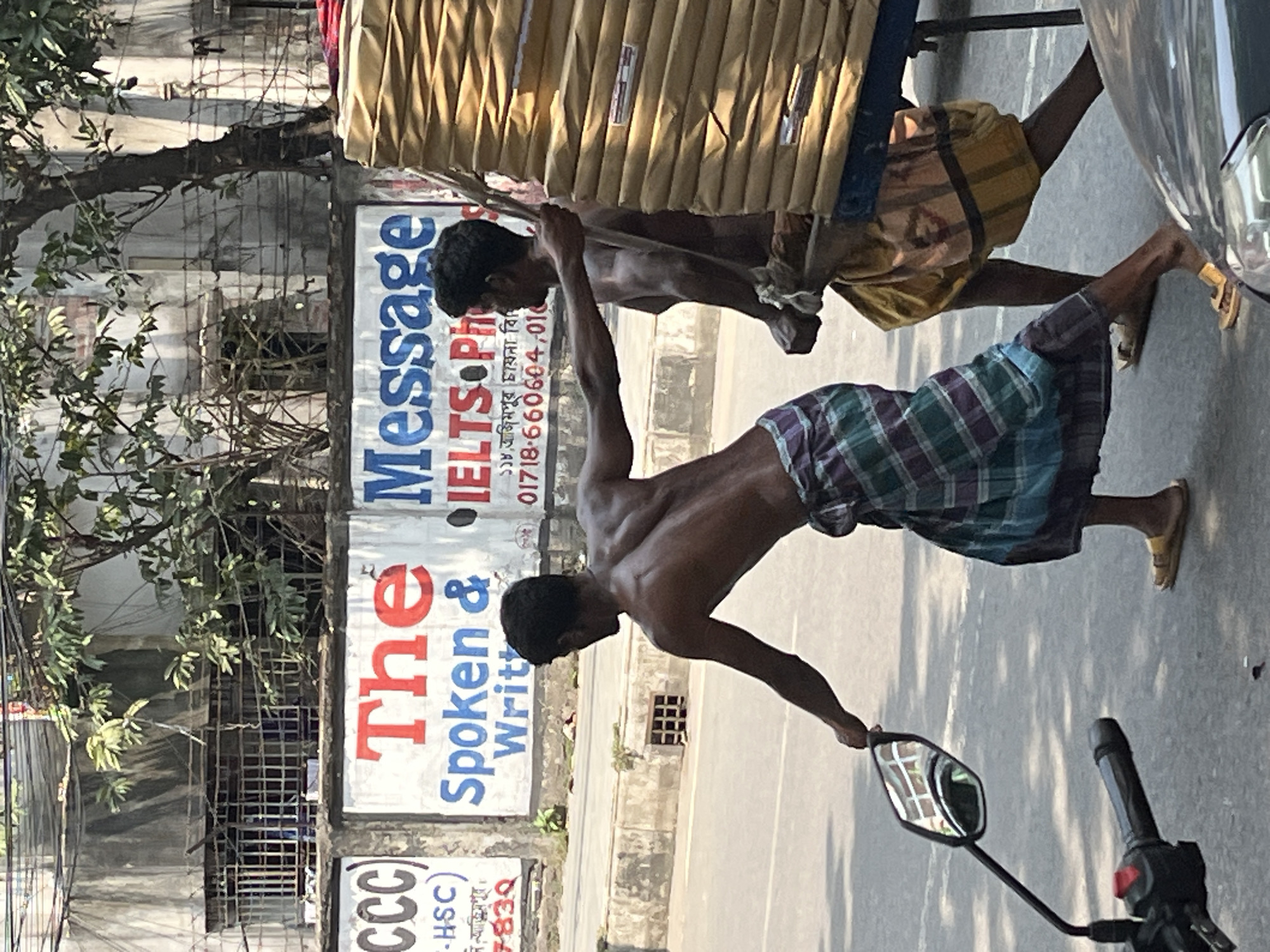 The image depicts two shirtless men in Dhaka, Bangladesh, transporting a large, wrapped bundle on a wheeled cart along a street. The men are wearing lungis, a traditional garment. One man is wearing a checkered lungi. The other man is wearing a striped lungi. The cart appears to be moving along the street. The street is paved. There is a motorcycle with a black handlebar and side mirror on the right side of the image. In the background is a building with signs for "The Message IELTS Photo." One of the signs mentions "Azimpur China Bhaban." There is also text with the phone numbers 01718-660604, 01. Additionally, there is a sign that says "CCC." The location is likely Azimpur in Dhaka. Trees are visible on the left side of the image. The cart and men are moving left to right in the image.