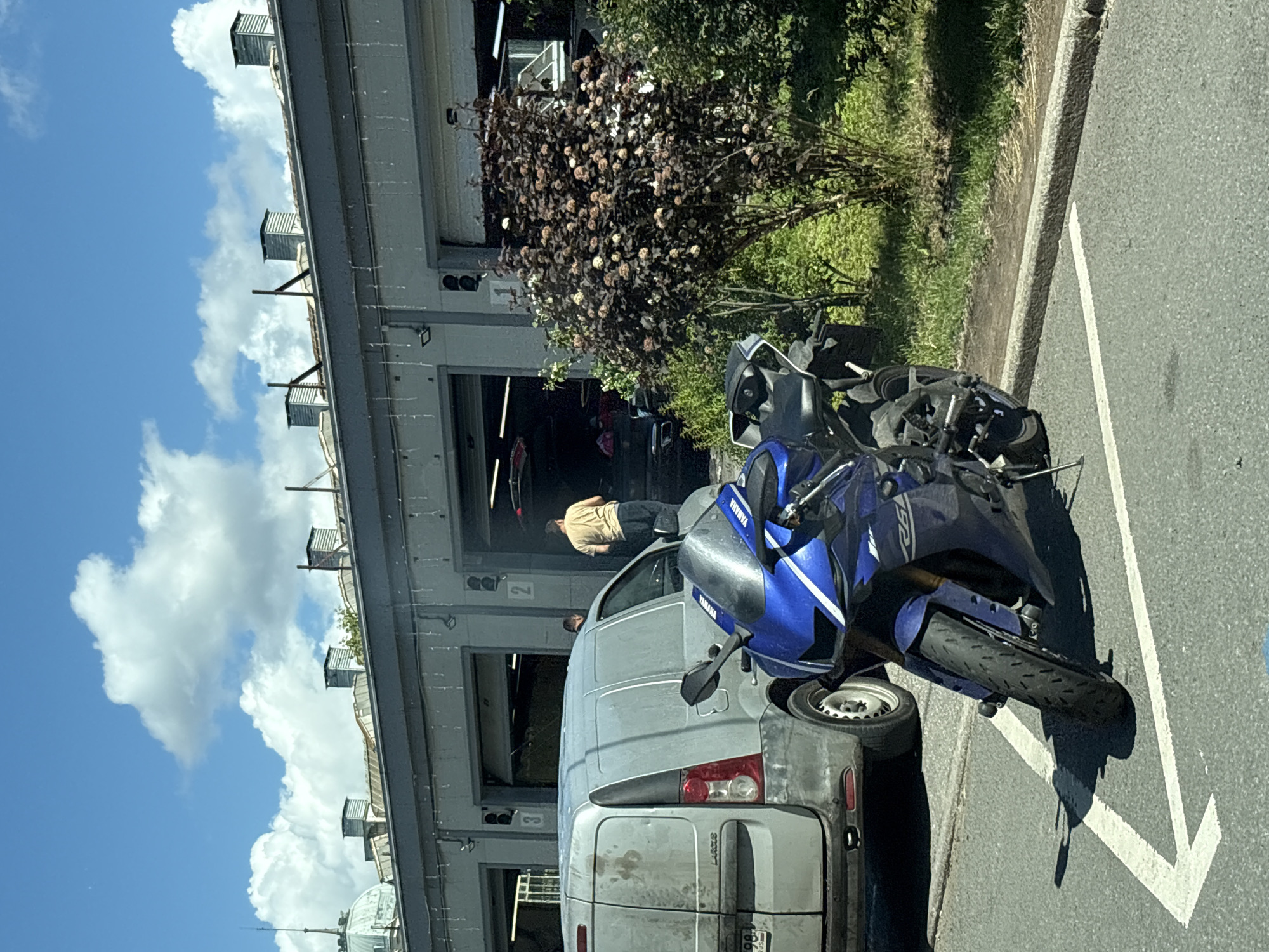 A sunny, partly cloudy day is captured in Saint Petersburg, Russia, outside what appears to be a garage or workshop with multiple open bays. In the foreground, a blue Yamaha sportbike is parked, its front wheel angled slightly to the left, casting a shadow on the asphalt. To the left of the motorcycle is the rear of a light gray Lada Largus van, its license plate showing Cyrillic characters and a Russian Federation flag. Behind the van, two men are visible near the garage bays. One man, wearing a tan shirt and dark pants, stands near bay number two, facing away from the camera and looking into the bay. Another man, whose face is not visible, stands near bay number three. A bush with dark leaves and small pinkish flowers is situated to the right of the garage bays, with green grass and more foliage behind it. The building itself has a row of identical garage doors, some of which are open, revealing interiors of vehicles. The roof of the building features several metal ventilation pipes and a historical dome structure visible in the far left of the image. The overall impression is a casual, functional urban scene on a bright day.