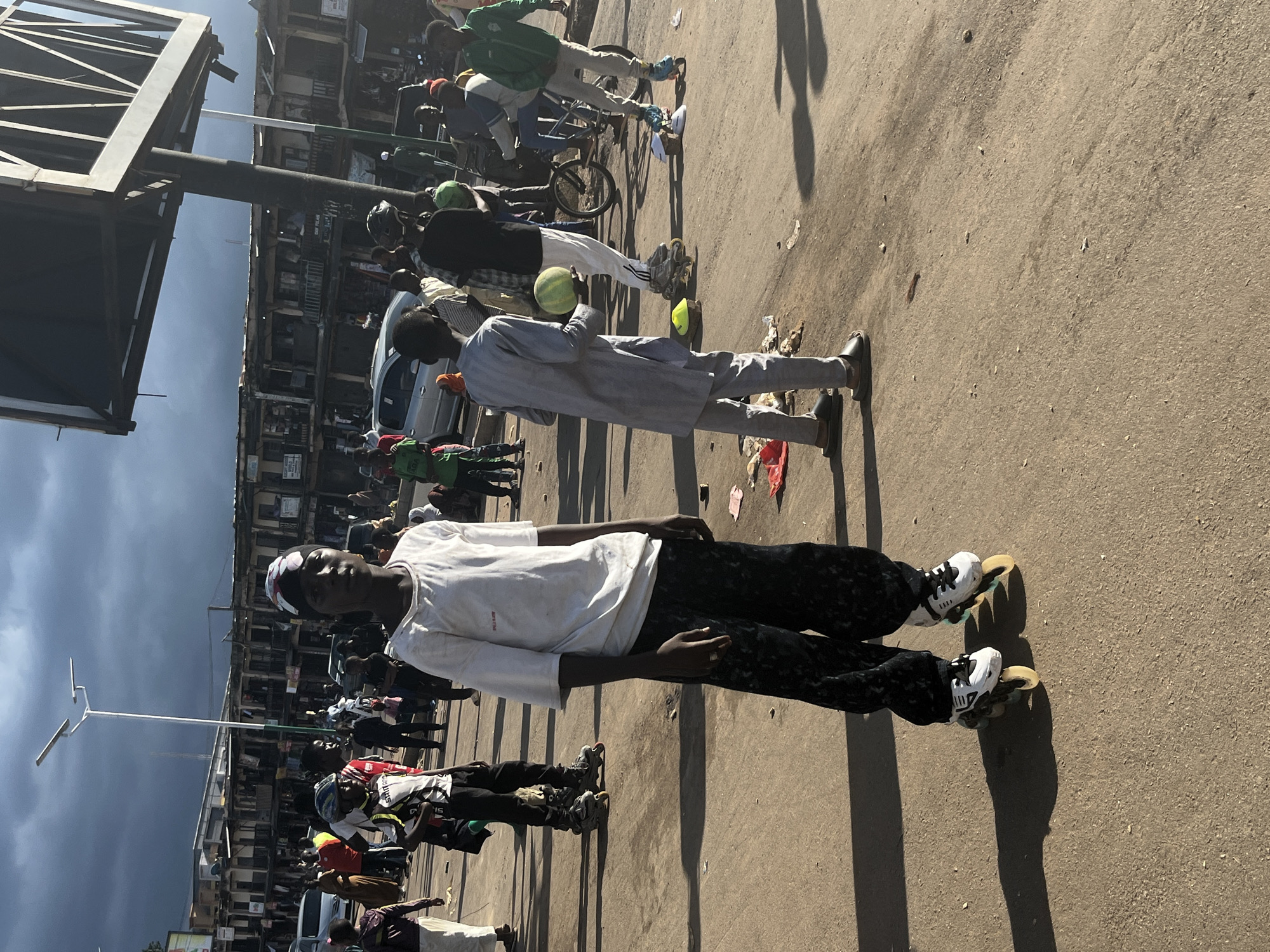 In Jos, Nigeria, on a cloudy day, a young person stands in the foreground on rollerblades, facing the camera. They are wearing a white t-shirt with a red logo and black patterned pants. Behind them, on a paved street, several other individuals are present, some on rollerblades, one holding a watermelon. In the background, a busy street scene unfolds with buildings lining one side and vehicles on the road. A tall lamppost and a large billboard structure are also visible. The ground is littered with some debris.