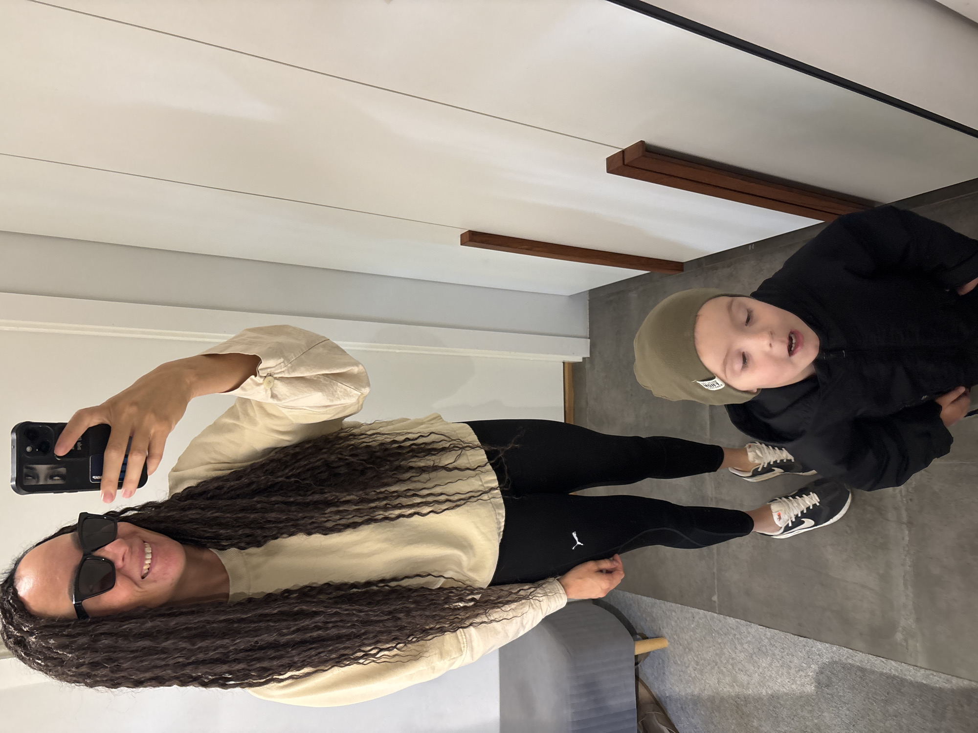 This indoor selfie, taken in Chaikovsky, Russia, features a woman and a young child. The woman, positioned on the left, has long, dark, textured hair or dreadlocks. She wears black sunglasses, a light beige long-sleeved top, and black leggings with a white Puma logo visible on her left thigh. She is smiling broadly and holding a black smartphone with a unique case displaying an image of eyes.
Beside her, on the right, stands a young child, likely a boy, dressed in a dark green beanie with partially visible text (possibly "FRONT"), a black padded jacket, and black pants. He wears black sneakers with white laces and soles, identifiable by a Nike logo. The child’s head is tilted slightly back, and his mouth is open, giving him an inquisitive or vocal expression as he looks upwards or slightly off-camera.
The background depicts a clean, modern interior space. The walls are white, and the floor is a light gray, possibly concrete. Decorative elements include horizontal wooden slats or beams on the upper wall and ceiling, alongside a minimalist track lighting system. A gray upholstered bench or seating unit is partially visible on the far left, suggesting a waiting area or fitting room. The lighting is bright, indicating an indoor setting.