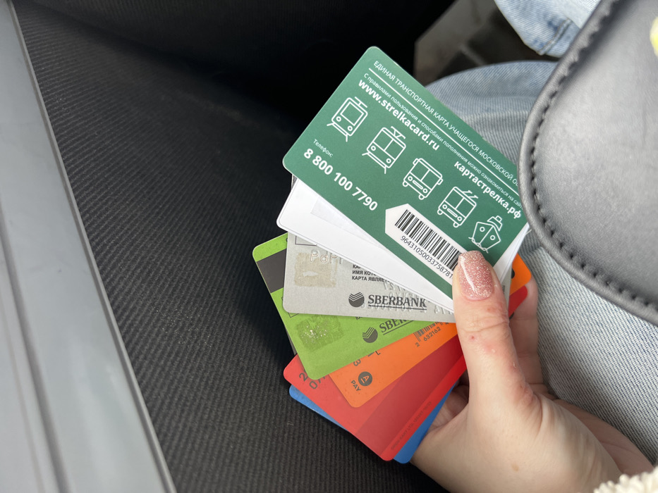A fair-skinned hand, likely female, with short, manicured nails painted with glittery pink polish, holds a fan of approximately eight plastic cards in the central foreground. Minor red bumps are visible on the thumb of the hand. The cards display various colors including blue, red, orange, green, grey, and white. Identifiable cards include a grey Sberbank card and a prominent green "Strelka" Unified Transport Card for Students of the Moscow Region. This green card features a barcode, the phone number 8 800 100 7790, the website www.strelkacard.ru, and icons representing various modes of transport. The individual's leg, clad in light blue denim jeans, is partially visible beneath the hand. A portion of a dark, stitched bag or purse is present in the lower right. The background consists of a dark, textured surface with horizontal ribbing, consistent with an interior panel, topped by a smooth, lighter grey stripe, suggesting an indoor environment, possibly within a vehicle. The image does not provide information to confirm the specific location within Lipitsy, Russia; rather, one card explicitly references the "Moscow Region."