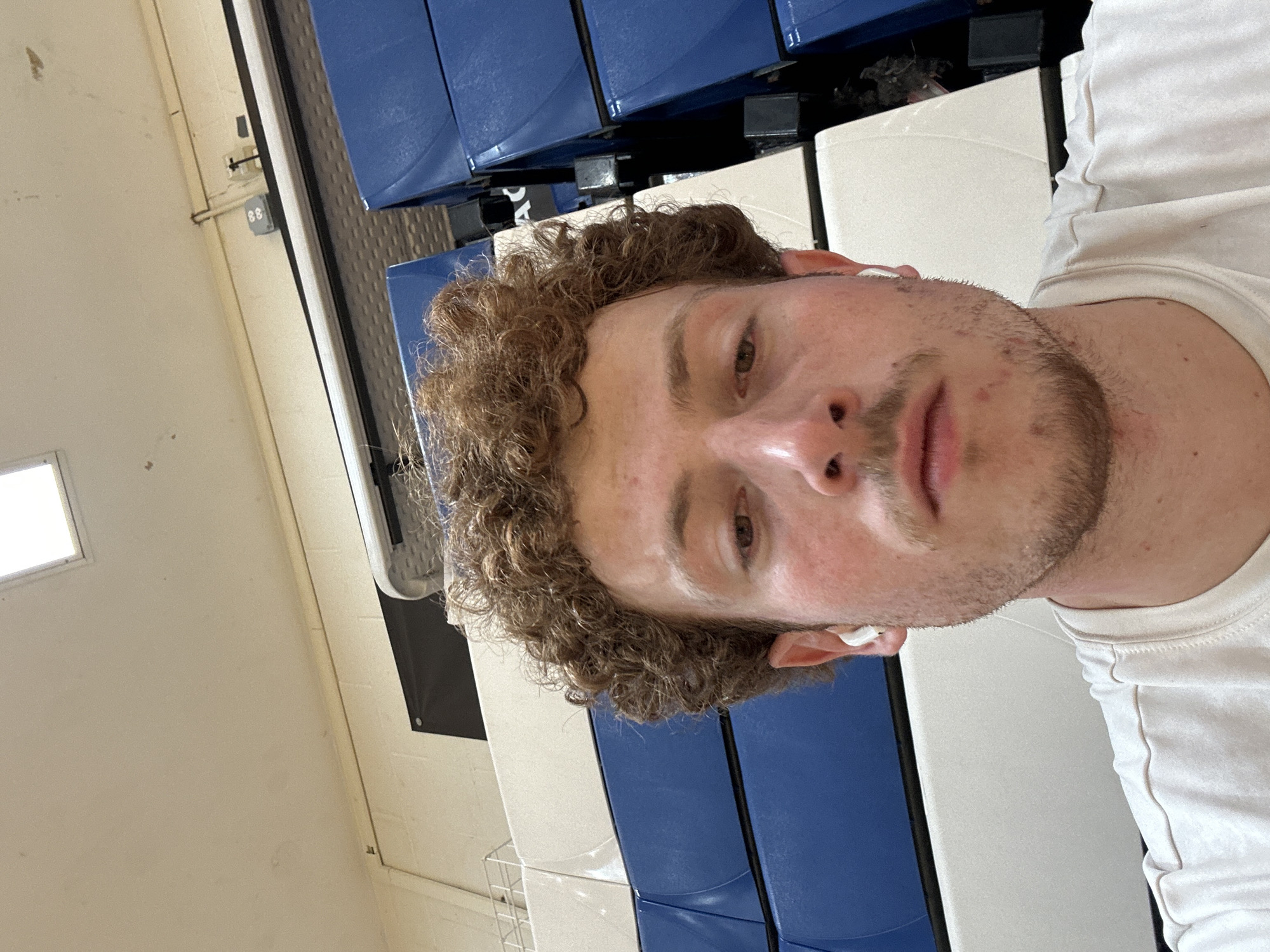 A selfie-style portrait features a young man with curly brown hair and light stubble on his chin and upper lip. He has a fair complexion and appears to be looking directly into the camera with a neutral expression. He is wearing a plain white t-shirt and white wireless earbuds in both ears.
The setting appears to be indoors, likely a gymnasium or sports facility. Behind him, visible mostly on the right side of the frame, are rows of blue padded seating, characteristic of bleachers. The background also includes light-colored walls, with a bright rectangular window visible in the upper left corner, suggesting natural light from outside. A small, grey box with the number "88" clearly visible on its surface is attached to the wall in the mid-left background. Other subtle details include a faint wire rack structure in the bottom left and some black structural elements associated with the seating. The overall lighting suggests it is daytime. The man's face fills a significant portion of the frame, indicating a close-up shot.