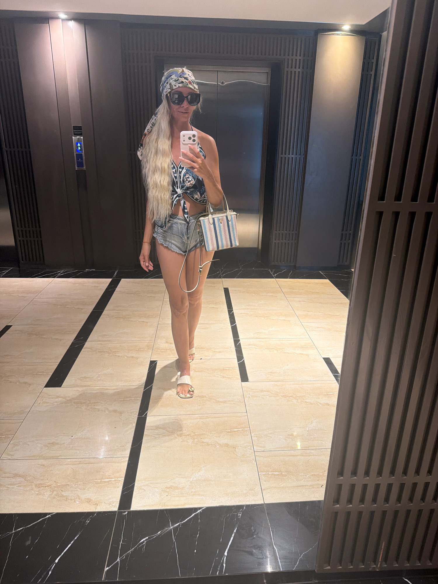 A blonde woman stands in a hotel lobby in Nha Trang, Vietnam, taking a mirror selfie. She is wearing a blue and white patterned halter top, denim shorts, a headscarf, and large sunglasses. She carries a small striped purse. The lobby has a dark wood paneled wall with an elevator and a floor with beige and black marble tiles.