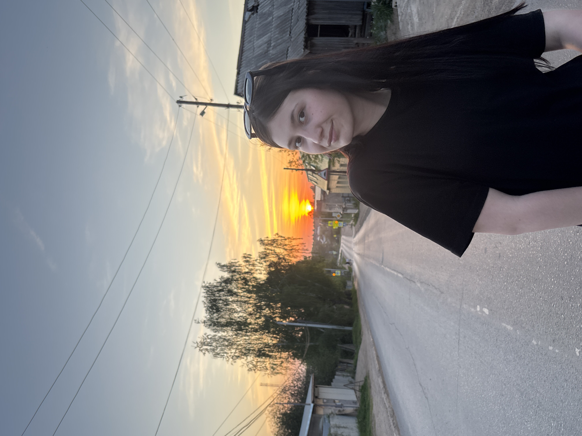 A young woman with dark hair and sunglasses perched on her head is posing for a photo on a street in Фоки, Russia. She's wearing a black t-shirt and standing on an asphalt road. In the background, a vibrant sunset illuminates the sky with orange and yellow hues. Trees line the street, and buildings are visible in the distance. Utility poles and wires stretch across the sky. An old, weathered wooden structure stands to the left of the woman.