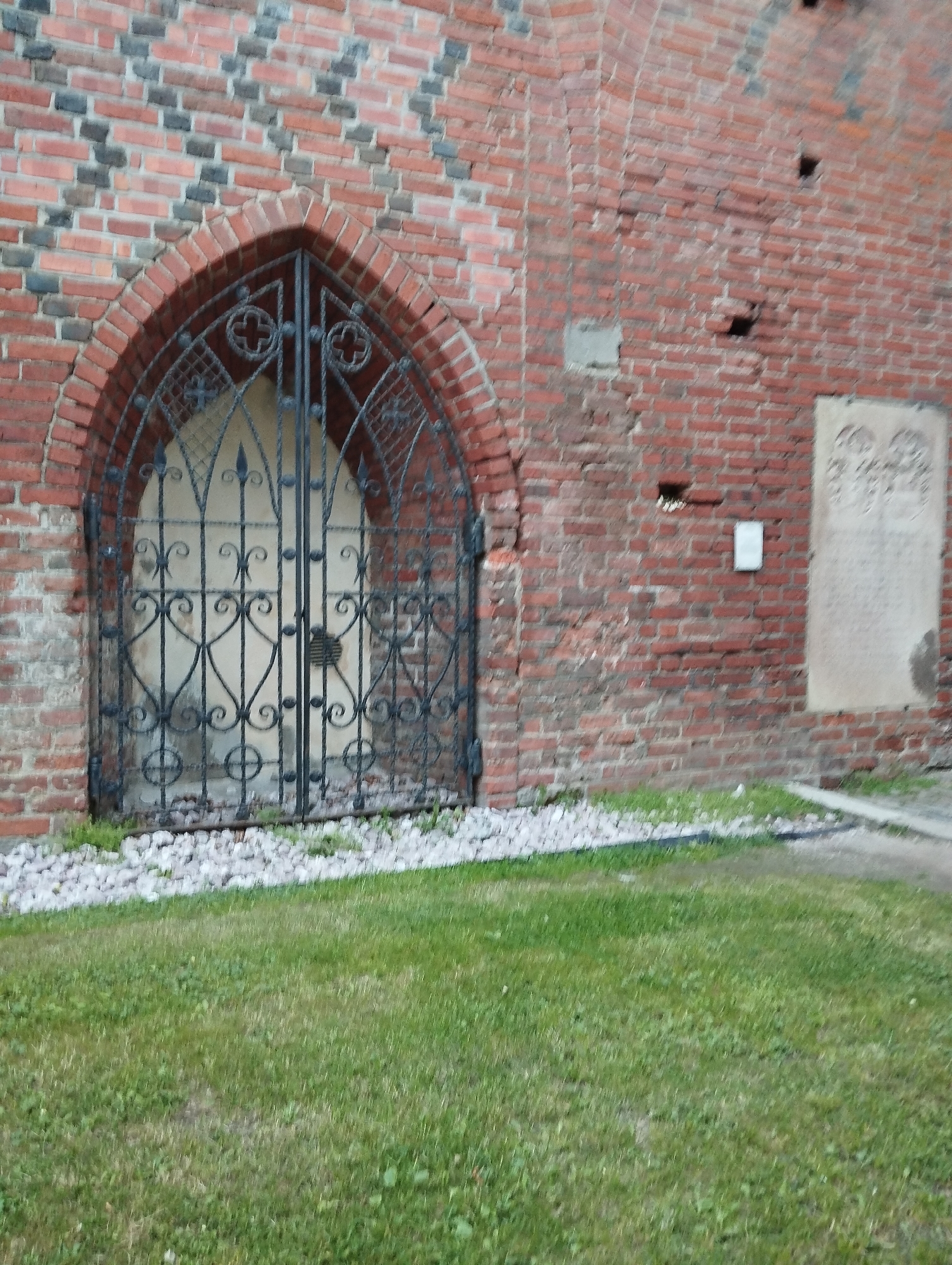 The image shows a close-up view of a weathered brick wall, characteristic of old European architecture. The wall has a repeating pattern of red and dark bricks. An arched opening in the wall is covered by an ornate, black wrought-iron gate with intricate scrollwork and decorative elements. The gate appears to lead into a dimly lit interior or alcove. In front of the gate, small white stones are scattered on the ground, bordered by a strip of green grass. The right side of the wall features a rectangular stone plaque with carved inscriptions, partially obscured by shadow. The overall scene suggests a historical or religious site. This location is likely part of the Königsberg Cathedral or one of its surrounding structures in Kaliningrad, Russia.