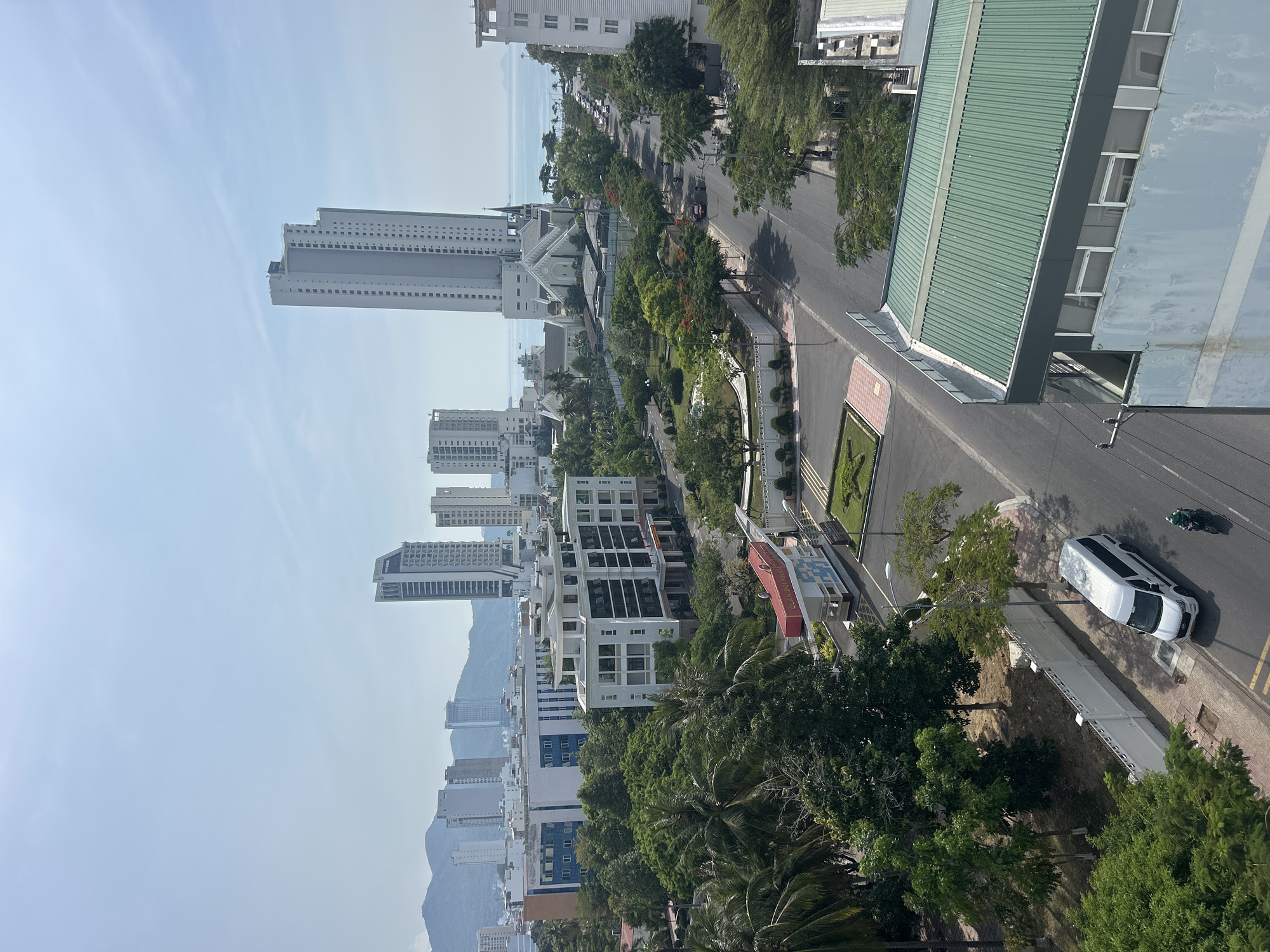 The image depicts a daytime cityscape in Bac Nha Trang Ward, Vietnam. A row of high-rise buildings, predominantly light in color, dominates the left side of the frame. Mountains are visible in the background behind the buildings. A prominent church structure is situated among the buildings closer to the center of the frame. A multi-lane road runs through the scene, partially obscured by trees. A white car is parked on the roadside. A green and white motorcycle is also on the road, heading the same direction as the white car. A bus shelter, partially red, is visible on the roadside. The foreground includes lush, green trees.