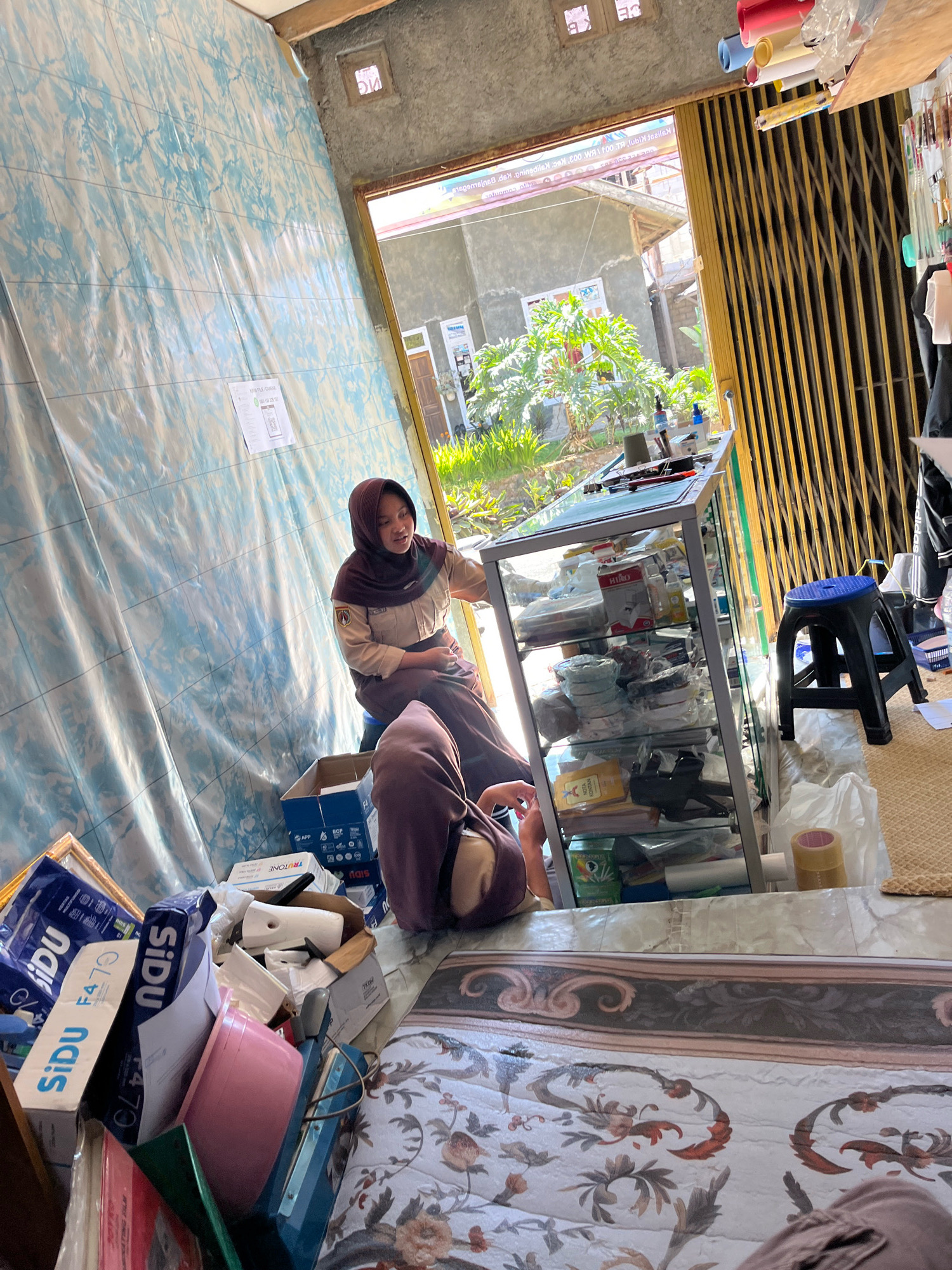 The image depicts an indoor scene, likely a shop or a storage area, in Krandengan, Indonesia. Two individuals, both wearing hijabs and modest clothing, are present. One person, positioned to the left and slightly further back, is seated on a low stool and appears to be looking towards the right. The other person, in the foreground and facing away from the camera, is seated on the floor.
The environment is cluttered with various items. To the left, there's a wall covered with a blue and white patterned plastic sheeting. A glass display cabinet filled with packaged goods is positioned in the center of the frame, extending towards the right. In front of the cabinet, there's a black plastic stool. Various boxes and plastic-wrapped items are piled on the floor, including what appear to be electronic components or small appliances, and a pink container. The floor is covered with a woven mat and a patterned fabric.
The background shows an open doorway leading outside, revealing a glimpse of vegetation and buildings, suggesting a residential or mixed-use area. A metal security gate is visible, partially obstructing the view outside. Rolls of colored paper or plastic are stacked on a shelf above the display cabinet. The overall impression is one of a busy, possibly disorganized, retail or storage space.