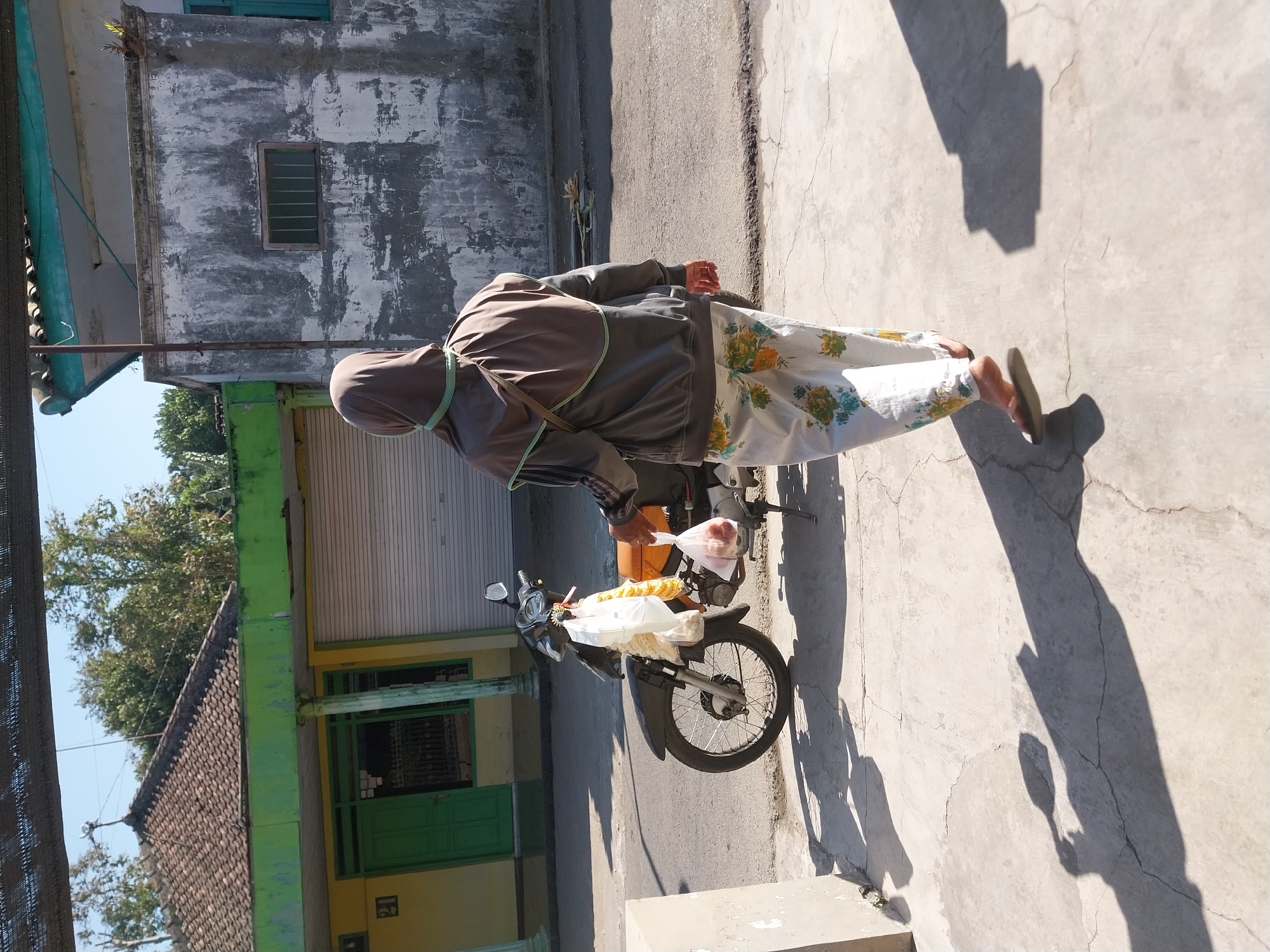A person wearing a brown hijab and a grey jacket with green trim walks away from the camera on a concrete sidewalk in front of a street. The person is also wearing white pants with a floral pattern and brown flip-flops. They are carrying a plastic bag in their right hand. To the left of the person is a parked motorcycle with items in plastic bags hanging from it. In the background, there is a building with a green and yellow facade and a tiled roof, as well as a grey building with a window. The scene appears to be in the city of Nglegok, Indonesia, indicated by the architecture and general setting. The ground is paved with concrete and shows cracks. Shadows are cast on the ground by the person and the motorcycle. The sky is clear and blue.