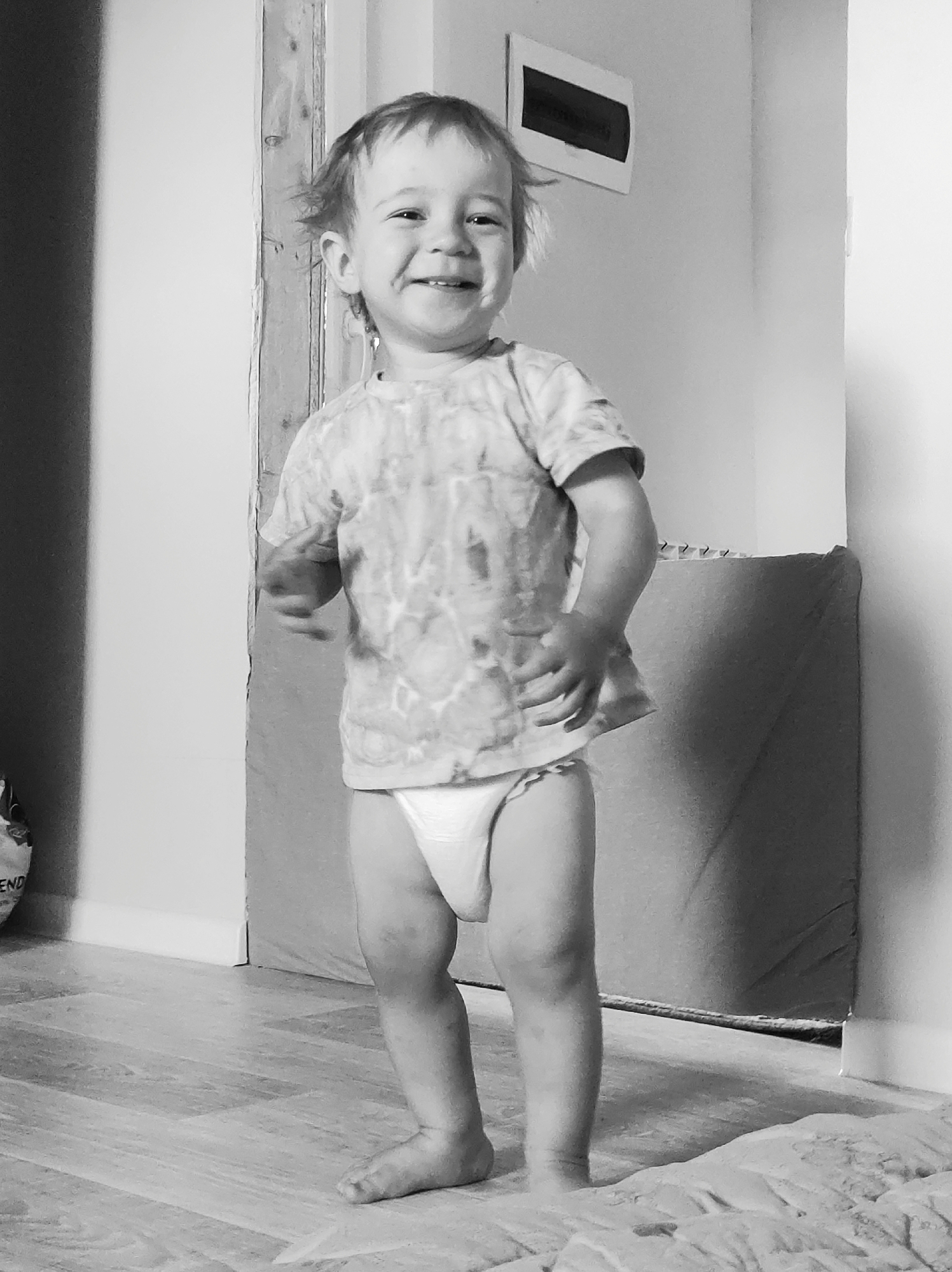A black and white, full-shot, eye-level photograph shows a young toddler standing in what appears to be a home environment. The toddler, facing the camera, has a wide, joyful smile and is wearing a t-shirt with a tie-dye pattern and a diaper. The child's hair is disheveled, and their skin appears fair. The background includes a wall with a framed rectangular object, a doorway, and a large, padded object. The floor is made of wooden planks. The lighting appears to be natural, coming from an unseen source. The overall impression is one of candid, everyday life.