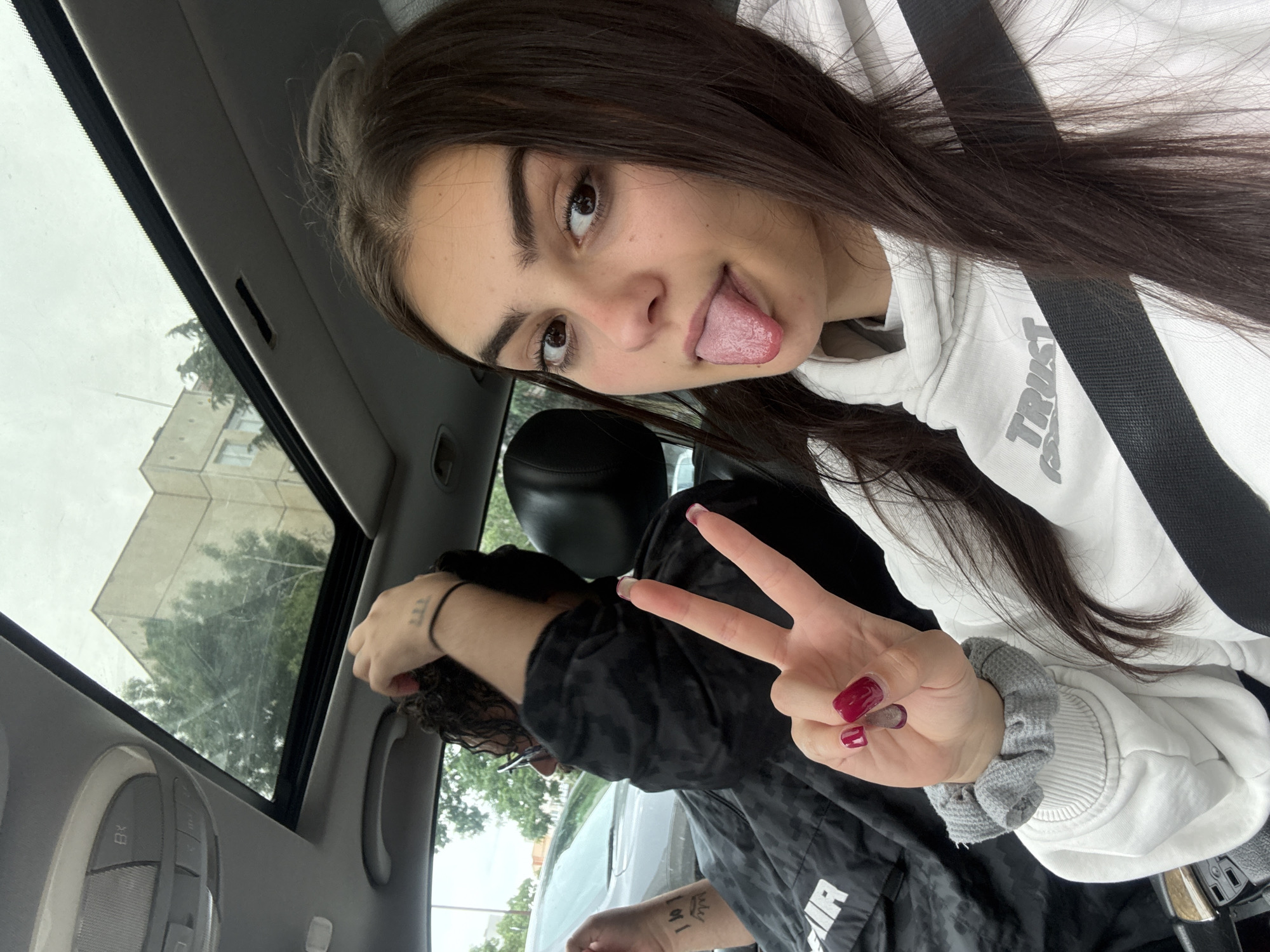 A young woman with dark hair and brown eyes is in a car, sticking out her tongue. She is wearing a white hoodie with the word "TRUST" printed on it. A black seatbelt crosses her torso. Her dark hair is parted to the side. Behind her, a person wearing a black jacket is making a peace sign with their hand. The person has dark curly hair, glasses, and a thin black bracelet on their wrist. Tattoos are visible on both hands of this person. Through the sunroof, a building and trees are visible. The car's interior is gray. The location is potentially in Blagoevgrad, Bulgaria, based on available information.