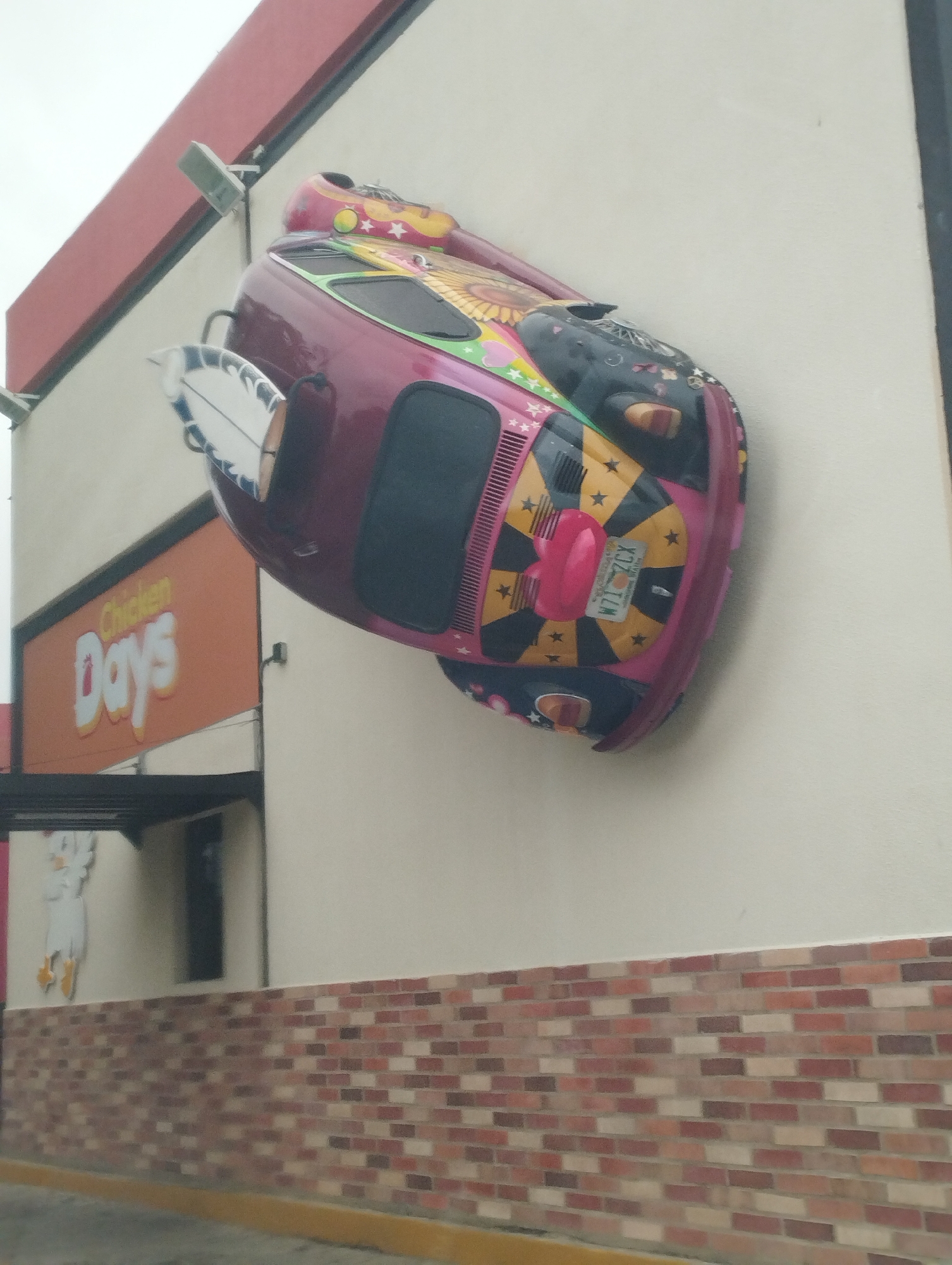 A colorfully decorated Volkswagen Beetle is mounted to the wall of a Chicken Days restaurant in El Tamarindo, Venezuela. The car, painted in pink, black, and yellow with various patterns, has a surfboard attached to the back. The restaurant is a single-story building with a red trim along the roof and a brick facade at the base of the wall. A sign for "Chicken Days" is visible on the building.