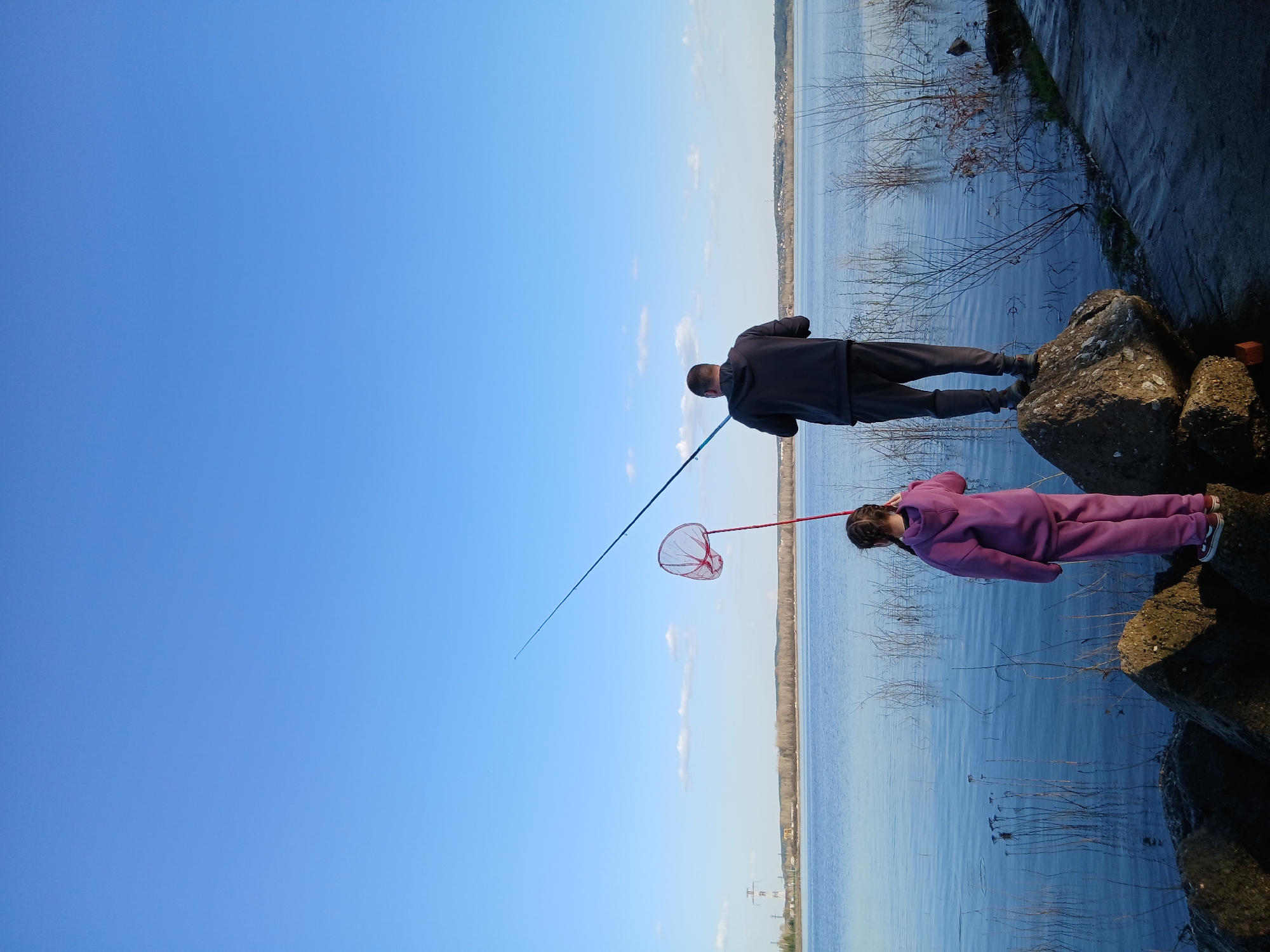 Two individuals are on the rocky shore of a large body of water, likely a reservoir or lake, located in Chaikovsky, Russia. One person, dressed in a dark tracksuit, is fishing with a long rod. The other, wearing a purple tracksuit, holds a red net. Both are standing on large rocks at the water's edge. The water stretches to the horizon, where there is evidence of distant buildings and land. Bare branches are visible in the water along the shoreline. The sky is clear and blue.