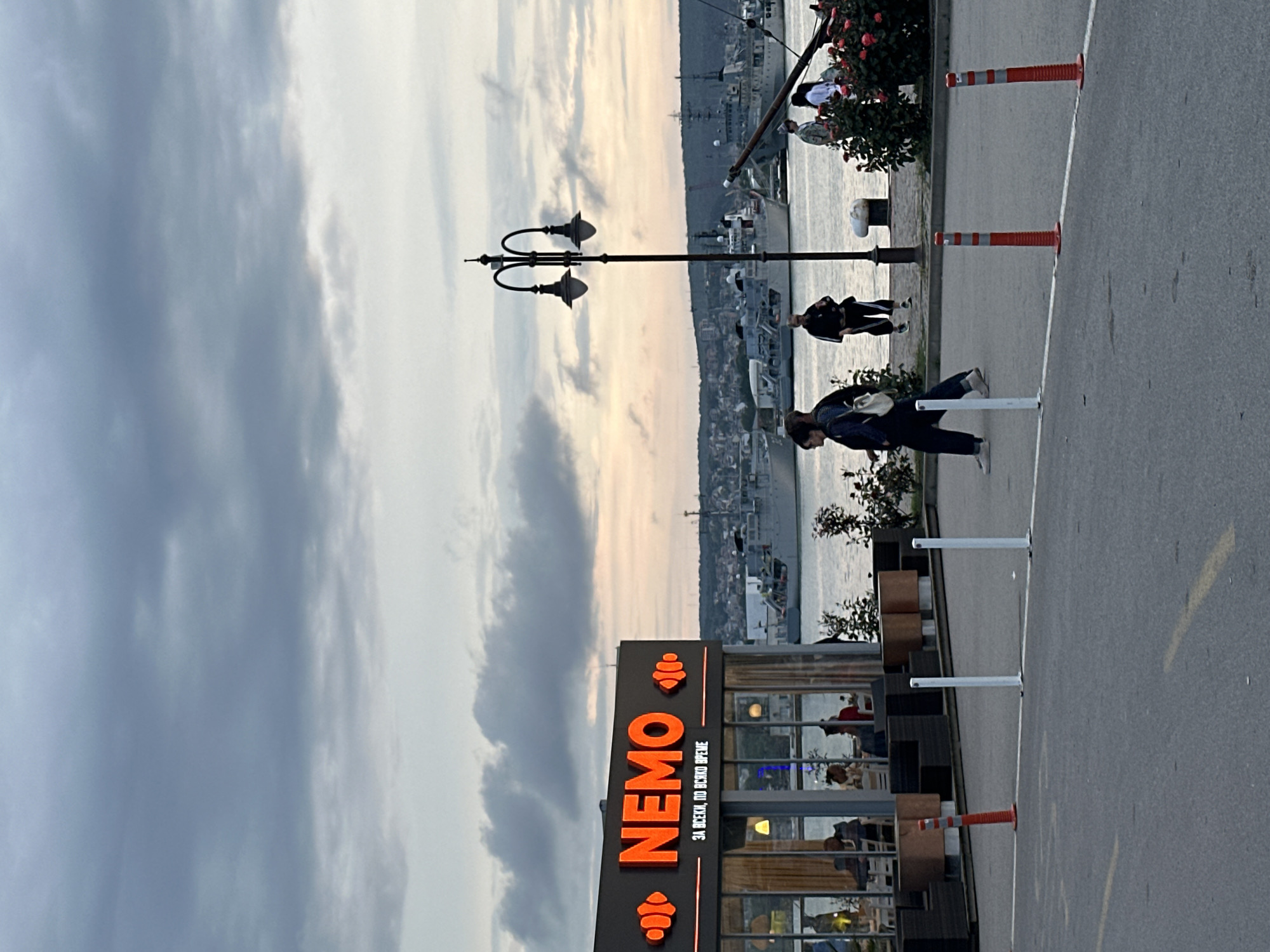 The image displays a waterfront scene, taken on an overcast day, likely during late afternoon or early evening. The perspective is from a paved area looking towards a body of water with a cityscape on the distant shore.
In the foreground, to the left, there is a building with a prominent orange sign that reads "NEMO" in large capital letters. Below "NEMO," in smaller Cyrillic script, is "ЗА ВСЕКИ, ПО ВСЯКО ВРЕМЕ," which translates to "FOR EVERYONE, AT ANY TIME." The building appears to be a cafe or restaurant, with large windows revealing people seated inside. Several dark-colored outdoor seating arrangements are visible in front of the building. White and orange bollards line the edge of the paved area, which could be a road or a promenade.
In the midground, a tall black street lamp with two lights stands out against the cloudy sky. Several individuals are present: one person is walking along the paved area in the middle-right, another is seated on a bench or low wall by the water, and a few others are near a rose bush by the waterfront. A distinct shoreline with buildings and what appears to be hills or mountains is visible across the water. Several boats or ships are anchored in the distance.
The sky is largely covered with clouds, creating soft, diffused lighting. The water reflects the sky, appearing calm. Given the user-provided context, the location is Varna, Bulgaria, a coastal city known for its Black Sea port. The overall mood of the scene is tranquil and leisurely.