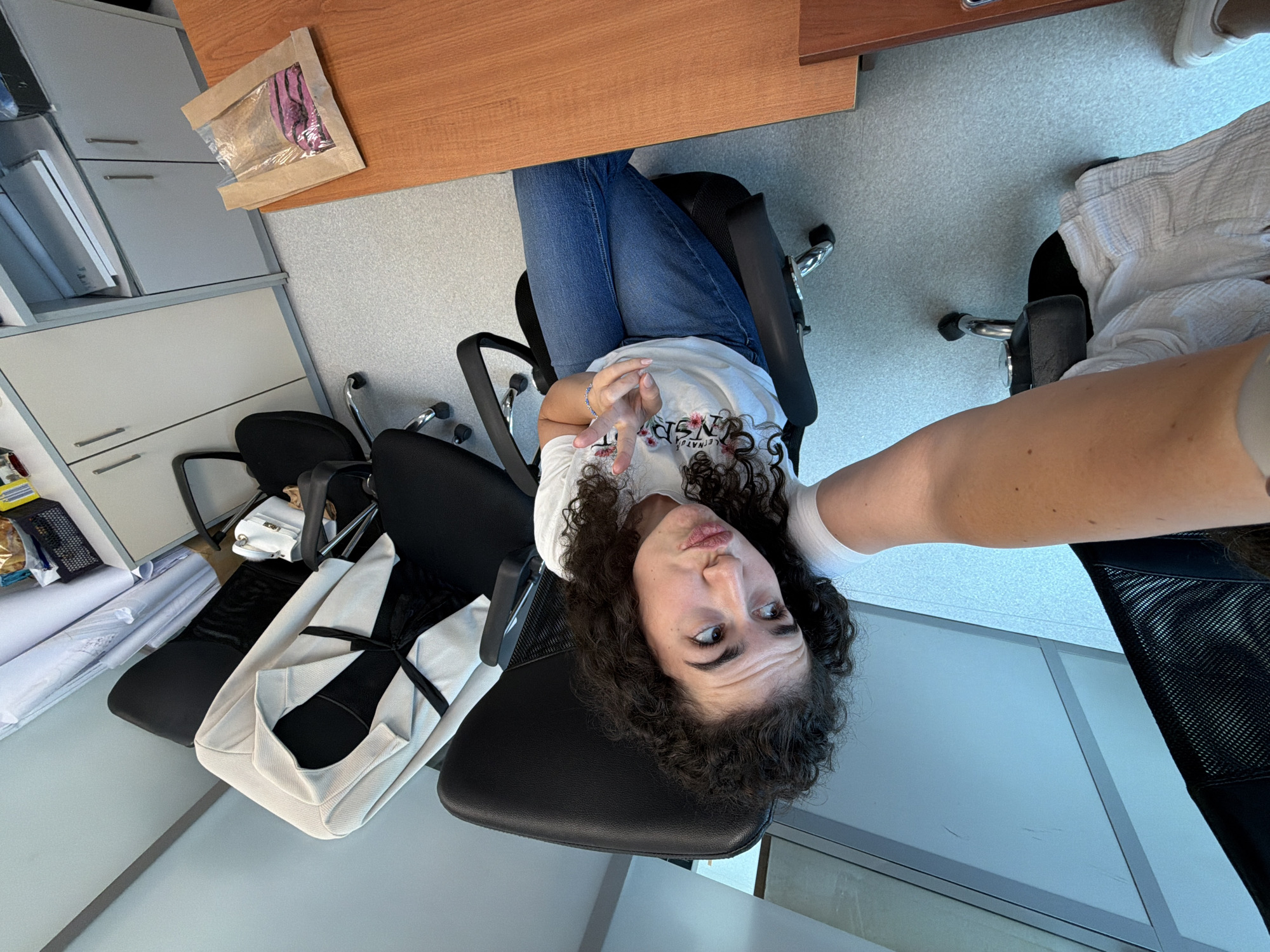 A person with dark, curly hair is sitting in a black office chair, facing the camera. They are wearing a white t-shirt with a floral print and blue jeans. The person is making a peace sign with their right hand. To their left, a light-colored blazer is draped over another black office chair. In the background, there are office cabinets and a wooden desk with a brown paper bag containing pink items. The scene appears to be an office environment.