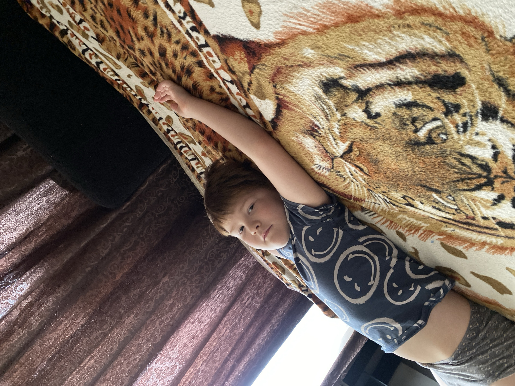 A young boy lies on a patterned bedspread, wearing a blue shirt with smiley face graphics and grey shorts. His arm is extended above his head. The bed is positioned near a window covered with brown curtains. A black object is seen next to the bed. The location is Chaikovsky, Russia.