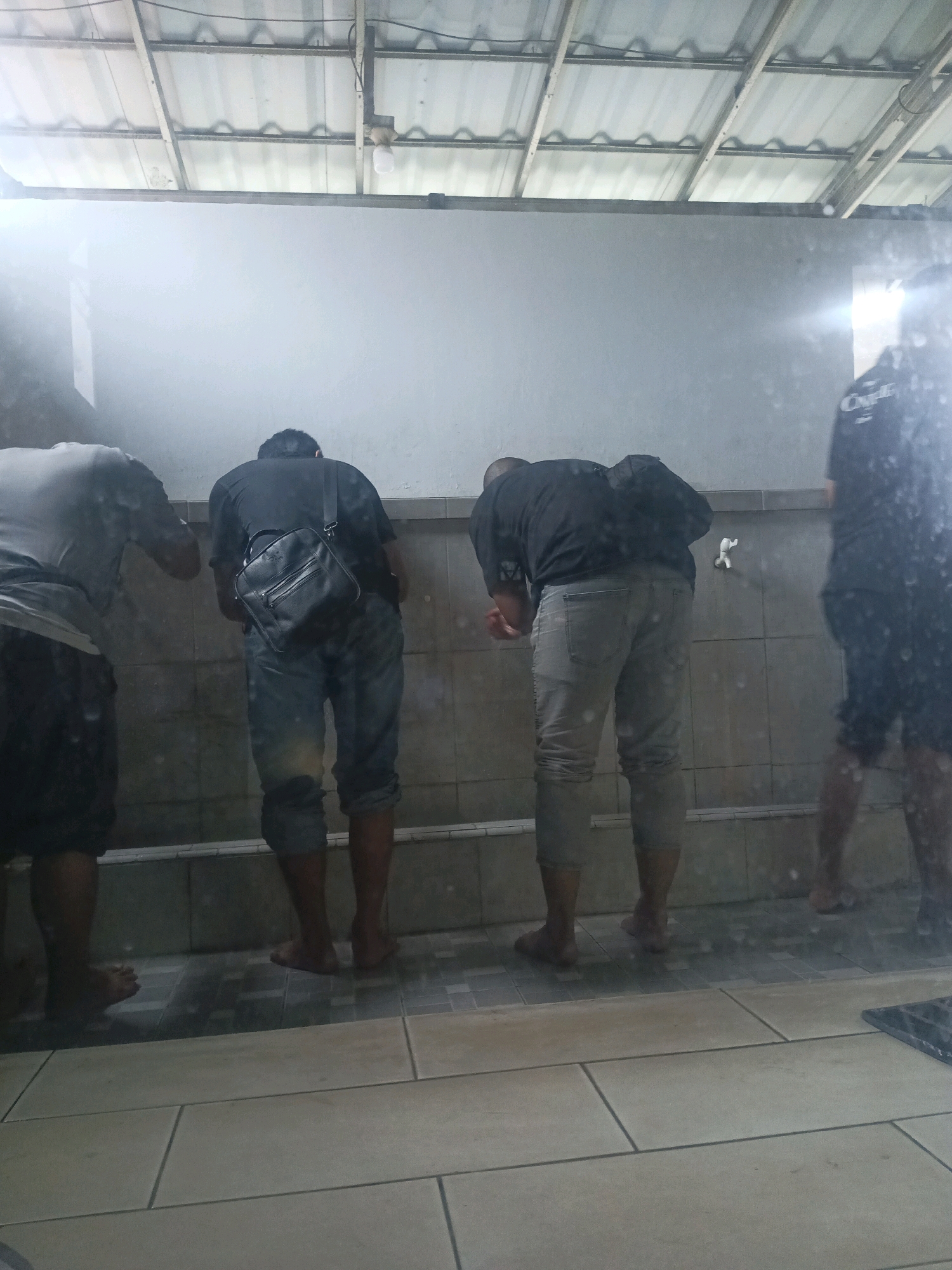 The image shows four men standing at a wash station, presumably a place for ritual ablution, as indicated by the presence of a faucet. The wash station is equipped with tiled walls and a trough at the base. The men are all facing the wall, with three of them appearing to be washing their hands or faces. The environment suggests an outdoor or semi-outdoor space, with a corrugated metal roof supported by metal beams visible overhead. The floor is tiled with large rectangular tiles. The scene is likely a public facility, possibly associated with a mosque or other religious site, located in South Tangerang, Indonesia. The lighting is somewhat dim, with a bright light source visible in the upper right portion of the image, creating reflections on a wet surface in the foreground. The overall impression is one of a functional, utilitarian space.