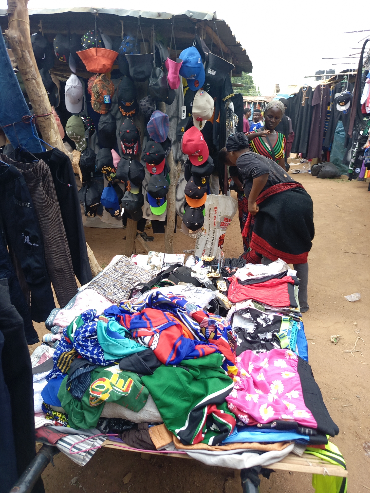 An outdoor market stall in Andaha, Nigeria, is depicted on a bright day. The scene features multiple items for sale, primarily clothing and headwear. In the foreground, a large table is laden with colorful piles of clothes, including t-shirts, shorts, and patterned fabrics. To the left, various dark trousers and jeans hang from a wooden pole. Above, numerous hats and caps of different styles and colors, such as baseball caps and bucket hats, are displayed, hanging from the roof of the makeshift stall.
Two women are prominently featured near the table of clothes. One woman, wearing a dark top and a black and red wrapper tied around her waist, is bent over, actively examining the merchandise on the table. The other woman, dressed in a vibrant, multi-striped top and a headscarf, stands upright, also looking at the items. In the background, other market stalls and a few additional people are visible, suggesting a bustling market environment. The ground is bare earth.
Visible text includes "LOVE." on a pair of dark pants, "HELLO" on a green shirt, "MO" and "M" on caps, and "UK Premium Pro 50KG NET," "GAMS," and "BIC DE Premium Milled Parboiled Ric" on a large white sack next to the bending woman.