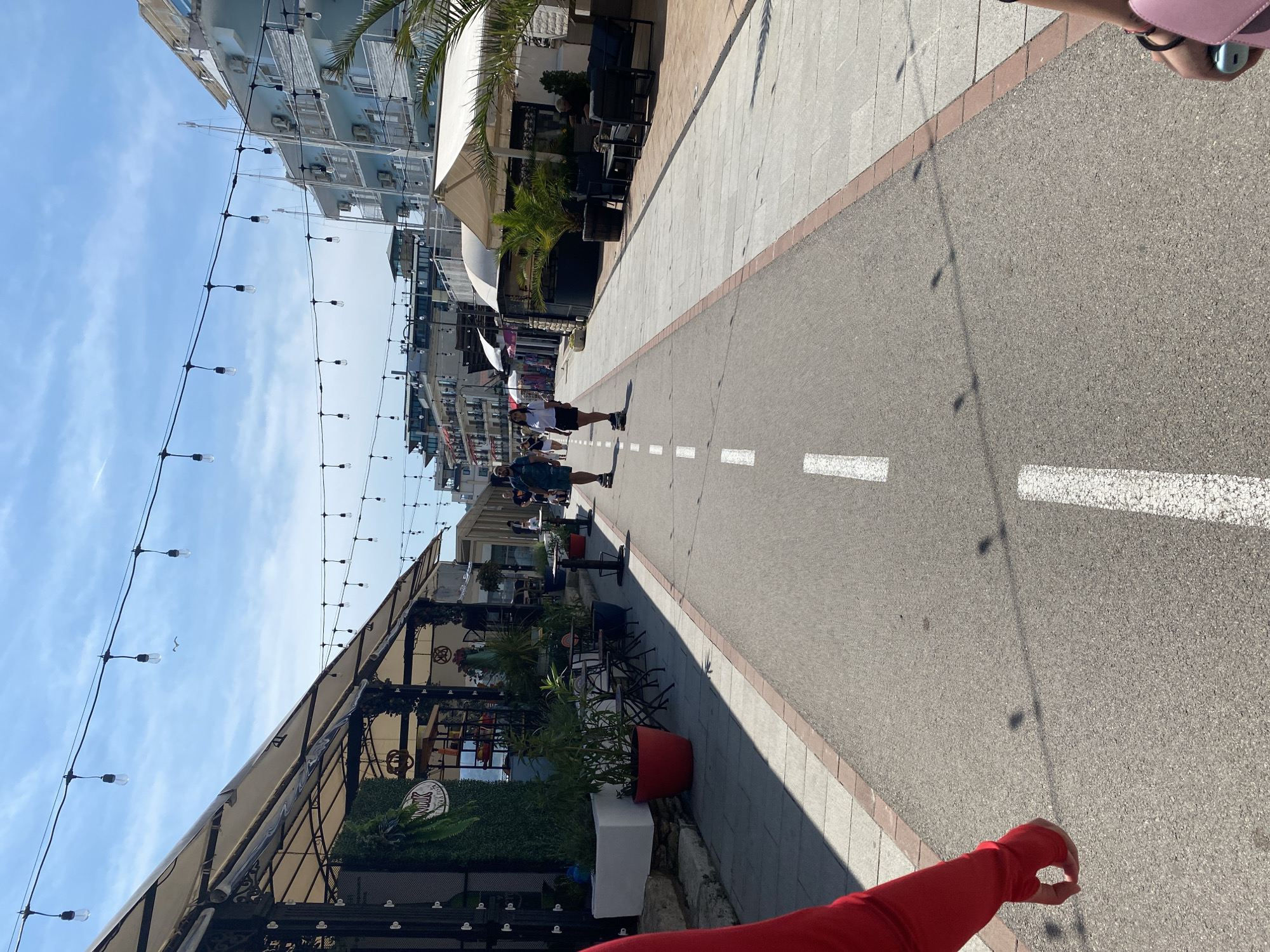 The image presents a sunlit commercial promenade in Balchik, Bulgaria, captured from a low, first-person perspective. A central asphalt path, marked by a dashed white line, extends into the distance, flanked by light-colored paved sidewalks bordered by reddish-brown bricks.
In the extreme foreground, the left side shows a partial view of a person's red-sleeved arm and open hand. On the right, another partial view reveals a hand holding a pink purse and a small blue object.
Along both sides of the promenade, numerous outdoor establishments, likely cafes or restaurants, operate under light-colored awnings. These establishments feature various elements:
* **Left side**: Potted plants, artificial greenery hedges, and outdoor seating comprising small tables and dark-framed chairs. One establishment displays a sign reading "MILK" followed by what appears to be "PIZZA PASTA."
* **Right side**: Similar outdoor seating arrangements, including lounge chairs, alongside potted palm trees. Further down, a prominent multi-story light blue building, likely a hotel, stands with multiple balconies and visible external air conditioning units. Other buildings are discernible in the background.
Two individuals are centrally located in the mid-ground, moving away from the viewer. Both appear to be riding skateboards. The person on the left is a male wearing a patterned shirt and dark shorts. The person on the right is a female wearing a white top and dark shorts. Other indistinct figures are visible further along the promenade and seated at the outdoor venues.
Overhead, multiple strands of string lights with bare bulbs are strung across the entire width of the promenade, casting distinct, elongated shadows onto the asphalt path. The sky is clear and bright blue with a few wispy white clouds, and a single bird is visible flying in the upper left section. The strong shadows cast by the lights and buildings indicate direct, strong sunlight, suggesting a midday scene.