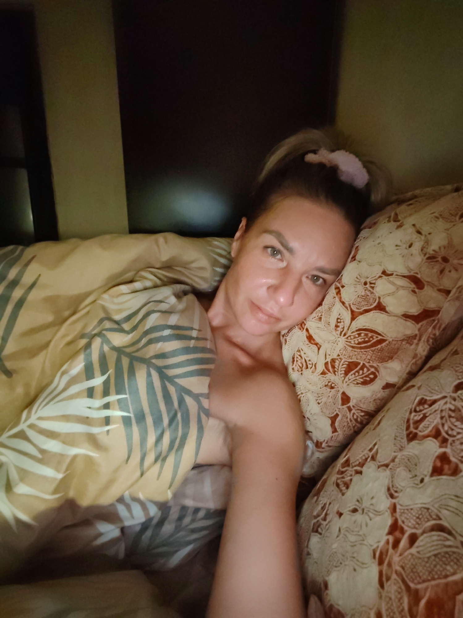 A woman is lying in bed. She is fair-skinned with light eyes. Her hair is pulled back into a ponytail with a pink scrunchie. She is resting on a floral-patterned pillow and covered with a comforter patterned with fern leaves. The setting appears to be a bedroom, with a dark headboard and bedside table visible. The image was most likely taken indoors and there is no indication of being in the city of Petropavlovsk-Kamchatsky, Russia.