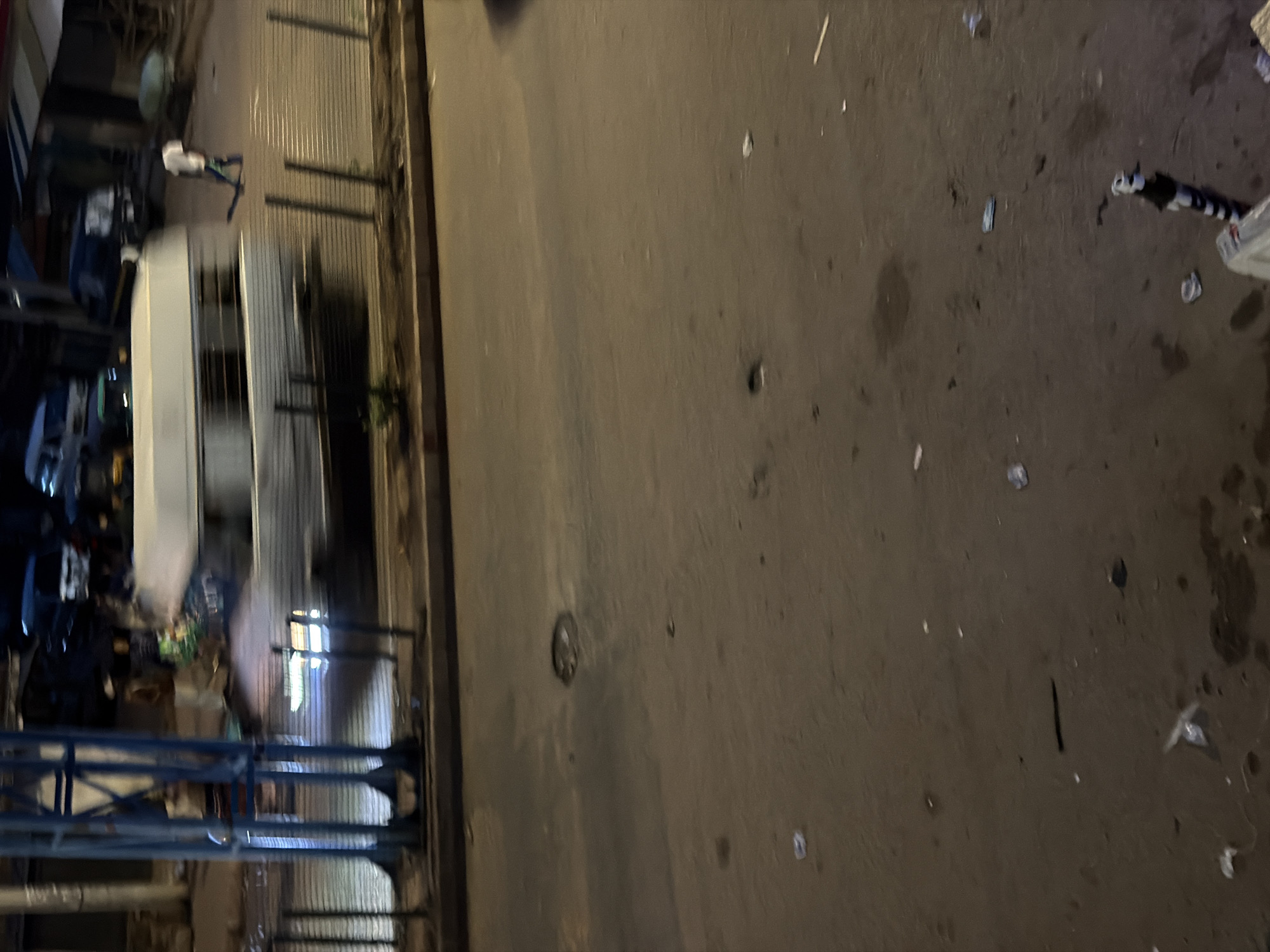 The image depicts a street scene at night in Bukuru, Nigeria. A white van is moving from left to right across the frame, captured with motion blur. A person in a white shirt and blue pants is walking away from the van, also blurred due to movement. The street surface is paved and appears somewhat worn, with some debris scattered on it. In the foreground, there's a metal pole with black and white stripes, and other objects that are difficult to discern due to the low light and motion. The background shows other vehicles and structures, but they are also out of focus and indistinct.