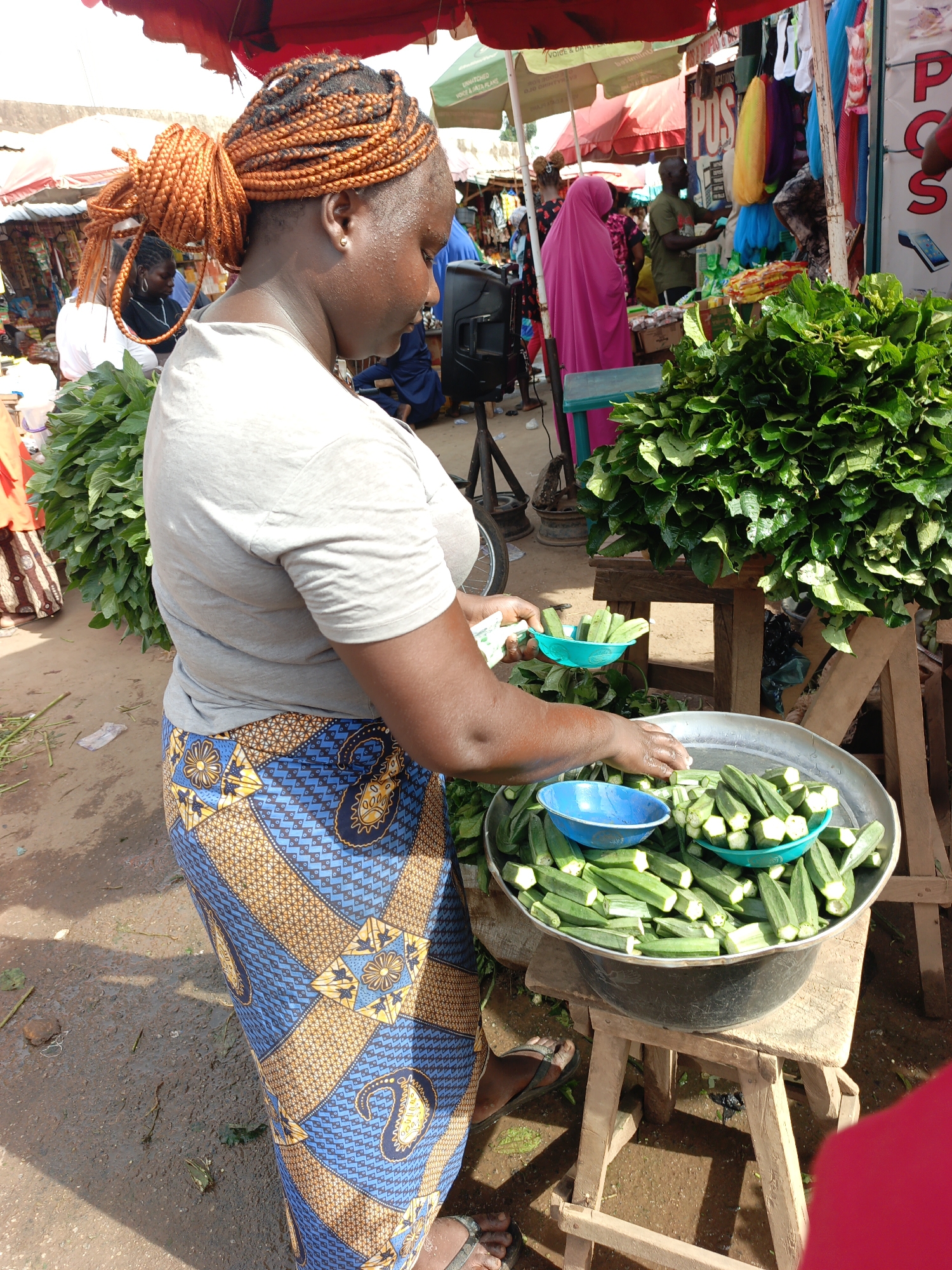 In a bustling marketplace in Akwanga, Nigeria, a woman with braided hair styled in a high bun, wearing a grey t-shirt and a blue and gold patterned skirt, is arranging okra in a large metal bowl atop a wooden stool. She holds a small teal bowl and is placing okra into it. Other teal bowls are scattered within the larger bowl. Beside her are bundles of leafy greens. In the background are various market stalls with people and colorful items under red and green umbrellas. A woman in a pink hijab is visible. A loudspeaker stands on a tripod in the periphery. The ground appears slightly muddy.