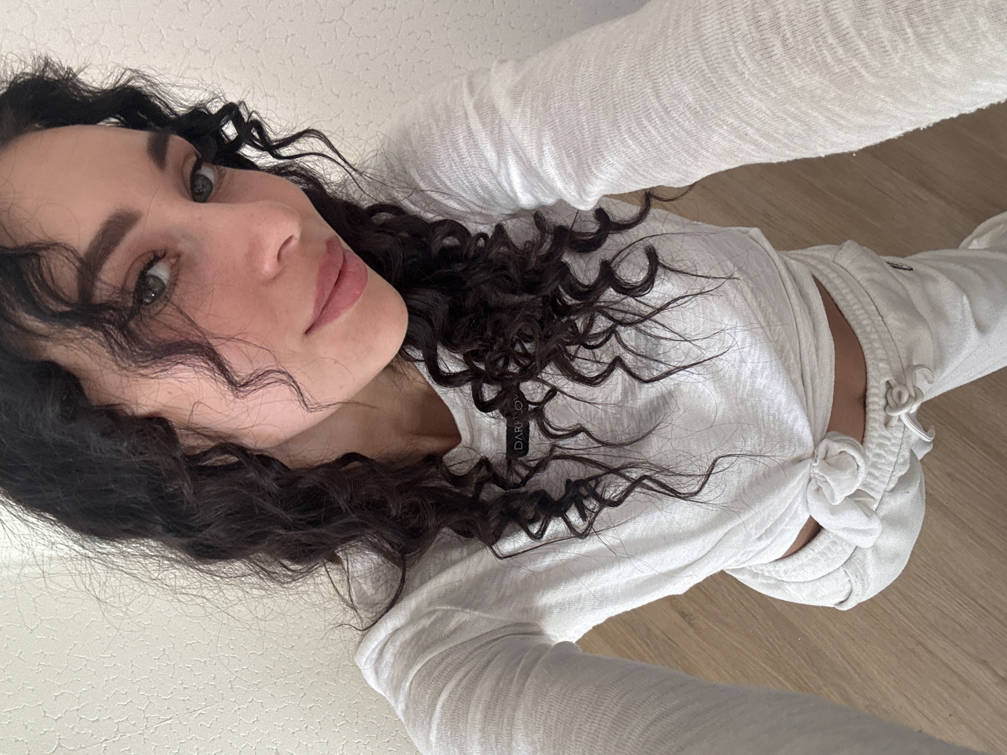 A young woman with long, dark, curly hair is captured in a top-down selfie, looking directly at the camera with a calm expression. Her head is tilted, and her light-colored eyes (appearing green or hazel) are prominent. She wears a white, long-sleeved top made of a textured, lightweight fabric, tied in a knot at her waist, revealing a sliver of her midriff. Light-colored, possibly white, casual pants or sweatpants complete her outfit. A black tag with the white text "DARK CLO" is visible on her top near her neckline. The setting is indoors, featuring a light-colored, textured surface (likely a ceiling or wall) and a light-toned wooden floor. The bright, even lighting suggests daytime or well-lit indoor conditions in Chaikovsky, Russia.