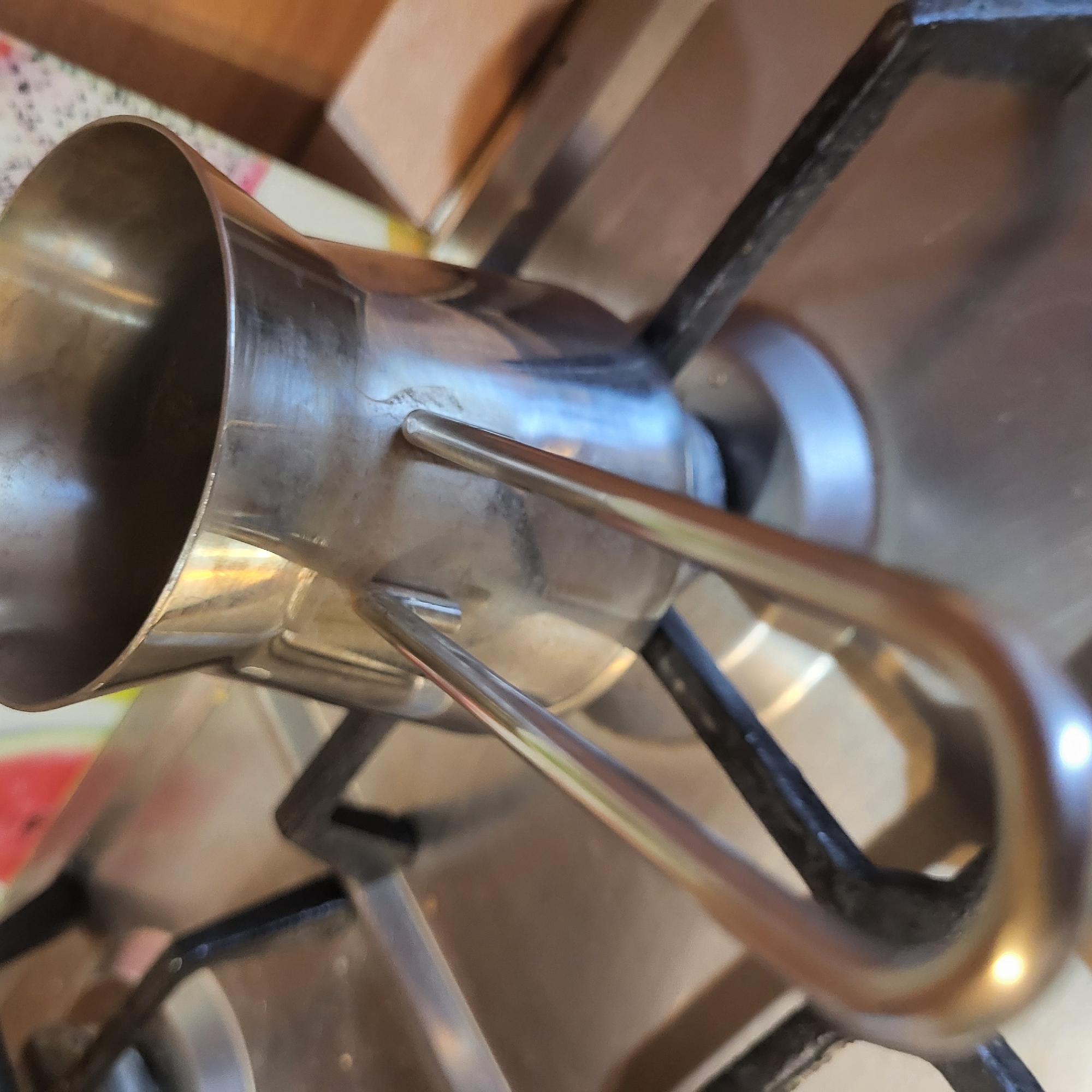 A close-up, angled shot captures a metal cezve (Armenian coffee pot) resting on the grates of a gas stove. The cezve, likely made of stainless steel, has a shiny, reflective surface that shows distorted reflections of its surroundings, including warm, blurry tones that suggest wooden cabinetry or a wall. Its design features a wider base tapering into a narrower neck, and then flaring out at the top rim. A long, slender, curved handle, also made of metal, extends from the side of the pot.
The cezve is positioned centrally over a burner opening on the dark metal grates of the stove, implying it is either about to be heated or has just been used for cooking. The stove grates appear sturdy and functional. The overall setting is an indoor kitchen, consistent with the preparation of coffee, a common practice in Yerevan, Armenia. No flame is visible, and the time of day and weather are not discernible. There is no visible text in the image.