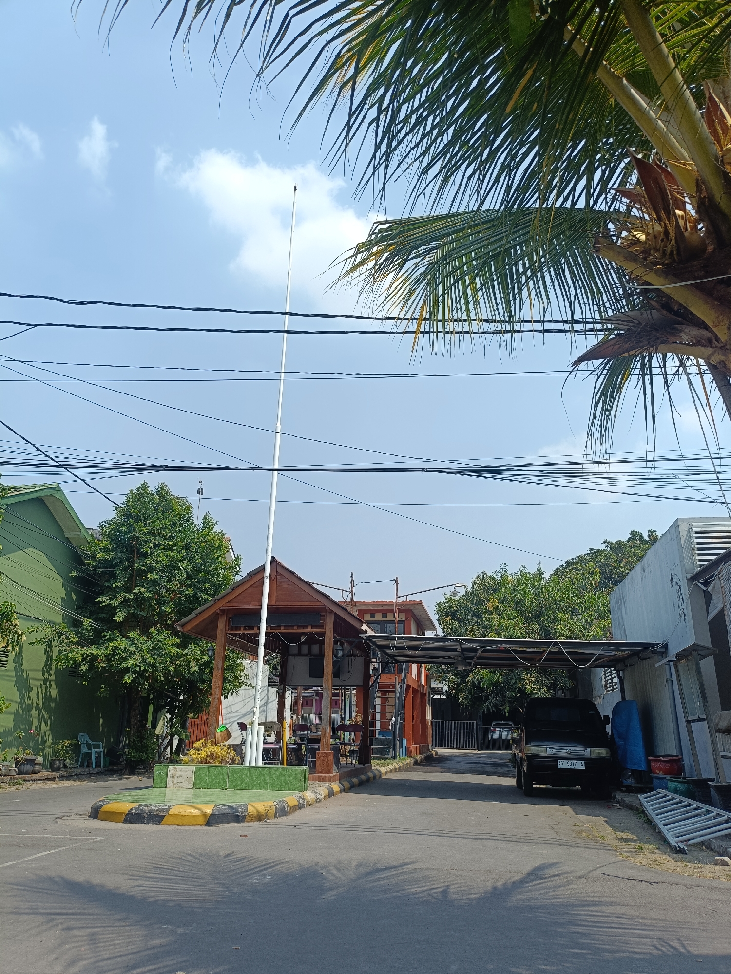 A wide shot captures a street scene in Kediri City, Indonesia, on a clear, sunny day. A tall, white flagpole stands near a wooden gazebo. In front of the gazebo, a raised platform is adorned with green tiles and bordered by yellow and black striped bollards. To the left, a green building stands next to a lush green tree. In the center of the image, a black pickup truck is parked under a metal awning. Further down the street, a large green tree is visible behind a red building, and power lines crisscross the sky. The foreground is dominated by an asphalt road with palm tree shadows cast across it.