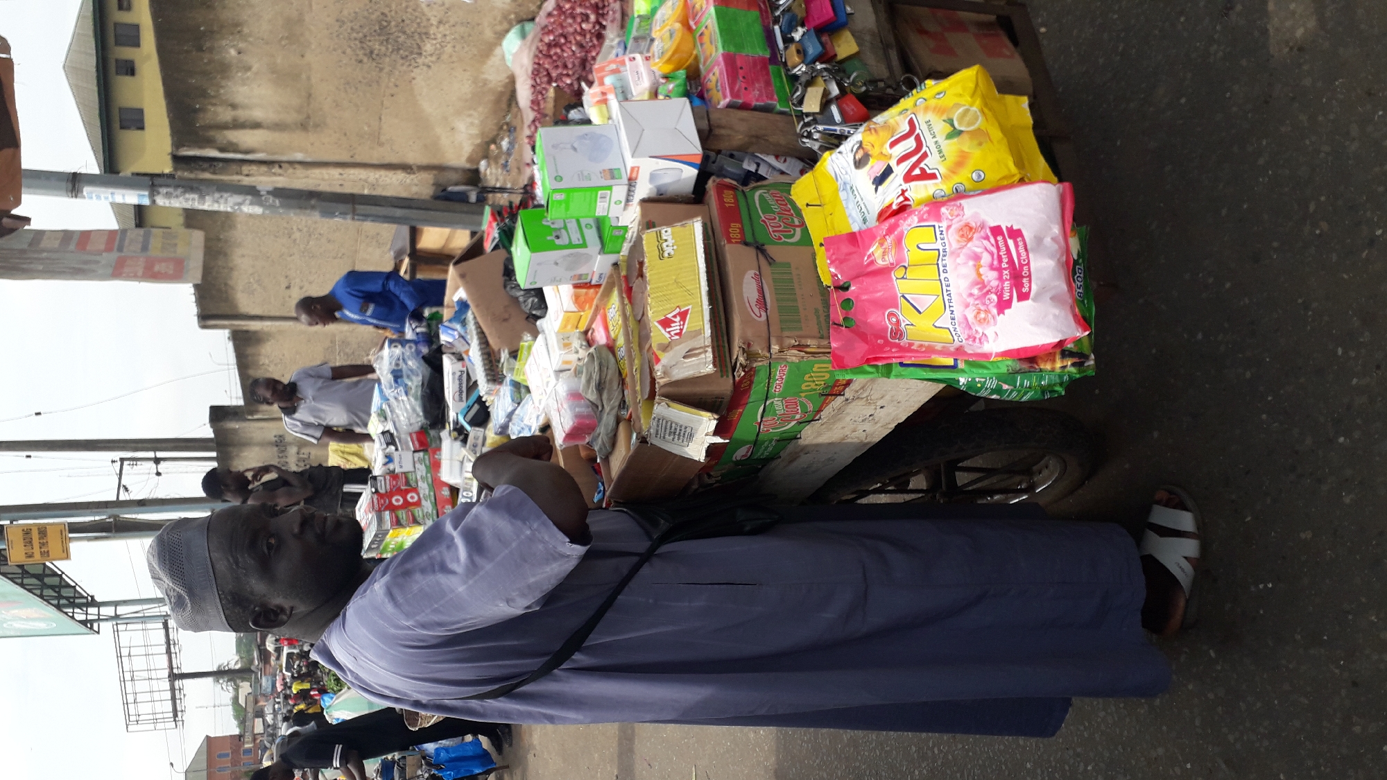 This image shows a street scene in Onitsha, Nigeria, likely a market. The foreground is dominated by a man wearing a purple robe and a skullcap, with a dark bag slung across his chest. His gaze is directed towards the left of the frame. Behind him, a variety of goods are displayed on a cart and on the ground, including boxes of detergent, soap, and other household items. Text visible on some of the packaging includes "ALL" (a detergent brand), "Attamuda", "CONCENTRATED DETERGENT", "With 2X Perfume", "Soft On Clothes", and "LEMON ACTIVE".
In the background, other individuals can be seen interacting around the stalls, including a man in a blue shirt and another in a grey shirt. The setting appears to be an outdoor market with a concrete wall and building in the distance. The lighting suggests it is daytime, possibly morning or afternoon, with overcast skies. The overall atmosphere appears to be one of commerce and daily life in a busy urban environment.