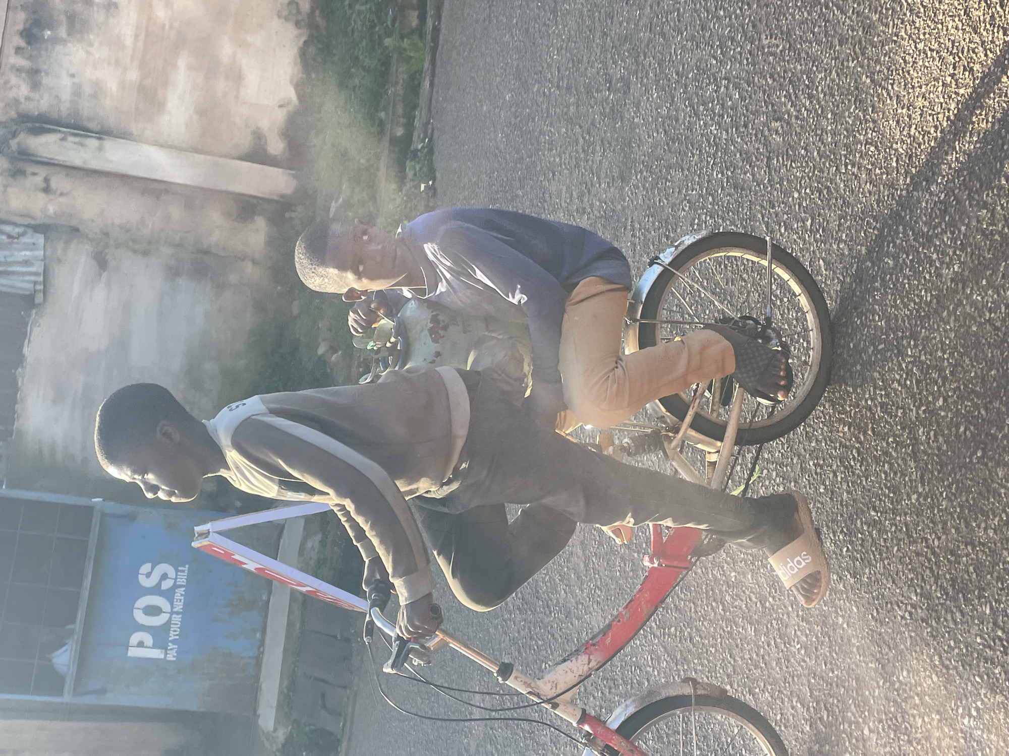 Two young boys are riding on a red and white bicycle on a paved road. The boy in front is pedaling and steering the bicycle. He is wearing a brown and white long-sleeved shirt and dark pants. He has Adidas slides on his feet. The boy behind him is sitting on the back of the bicycle. He is wearing a blue long-sleeved shirt and tan pants. He is holding onto a light green gas cylinder. They are in an outdoor environment, with a building in the background that has "POS PAY YOUR NEPA BILL" painted on it. The sun is shining, casting shadows on the road. The overall scene appears to be in the city of Jos, Nigeria.